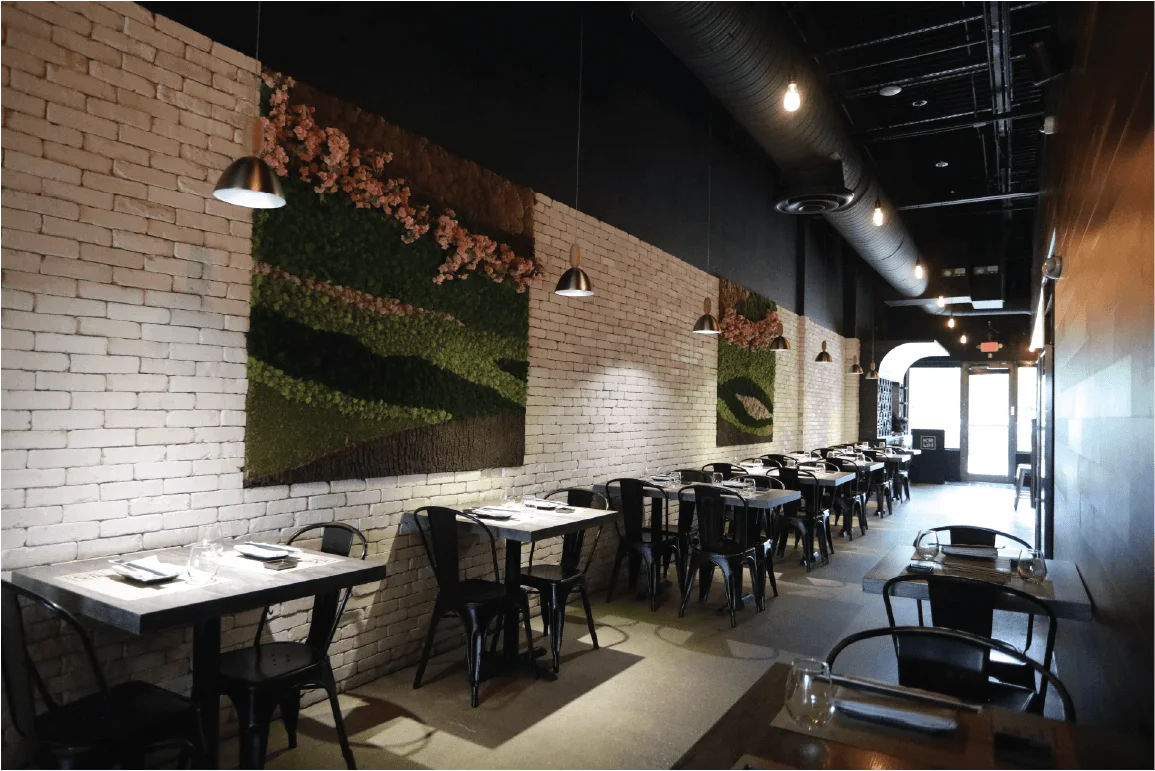 A restaurant dining area featuring a row of black tables and chairs along a white brick wall, decorated with moss wall art and industrial pendant lighting.