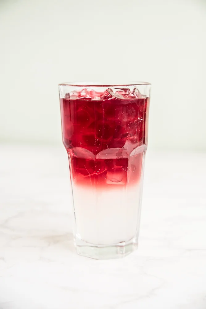A tall glass filled with a refreshing layered beverage. The bottom is creamy white, transitioning smoothly to a rich red on top, over ice cubes.