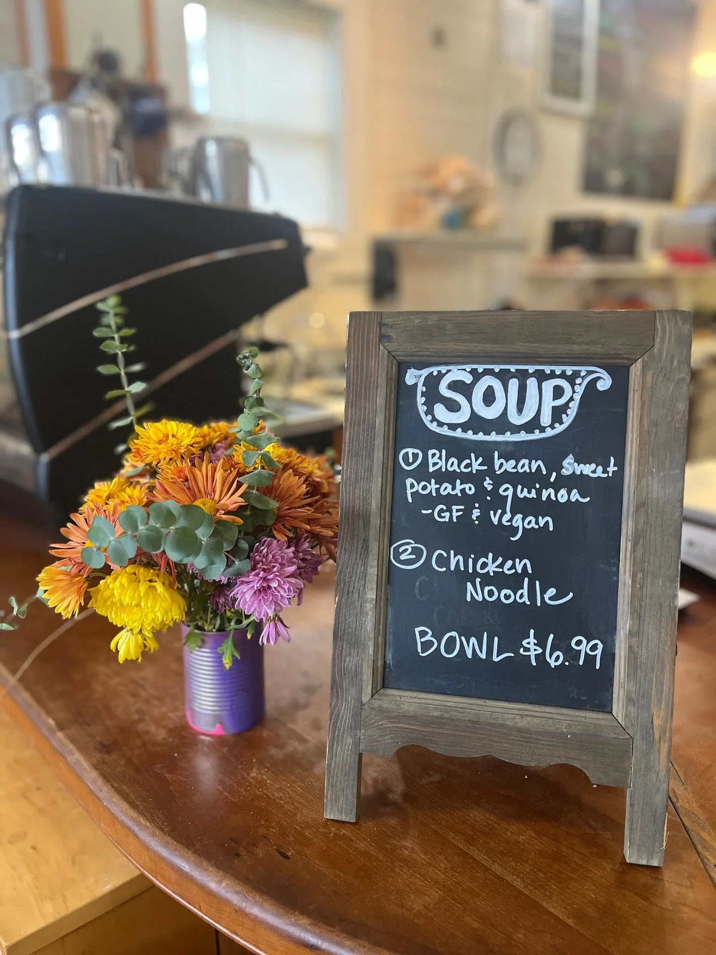 A chalkboard sign displaying two types of soup offerings - Black bean, sweet potato & quinoa (GF & vegan) and Chicken Noodle, placed beside a colorful floral arrangement.