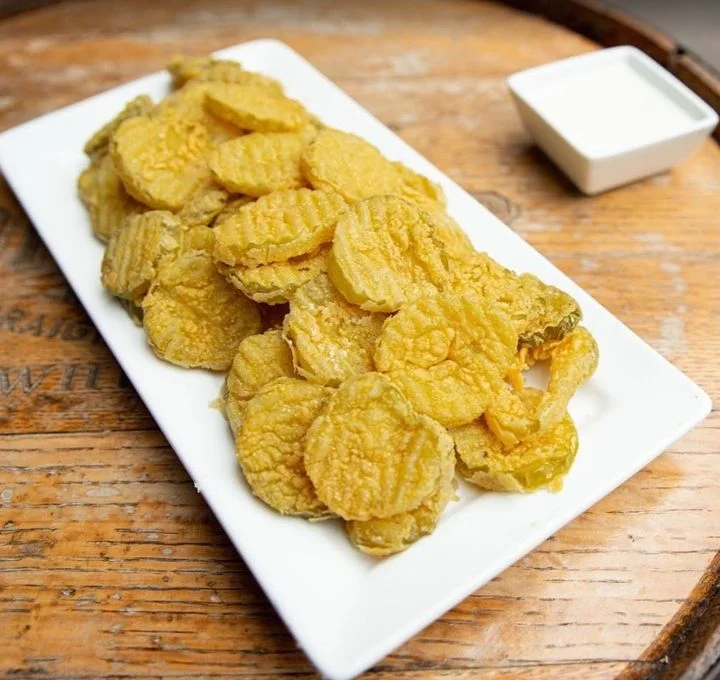 A plate of crispy fried pickles arranged on a white platter, accompanied by a small dish of dipping sauce.