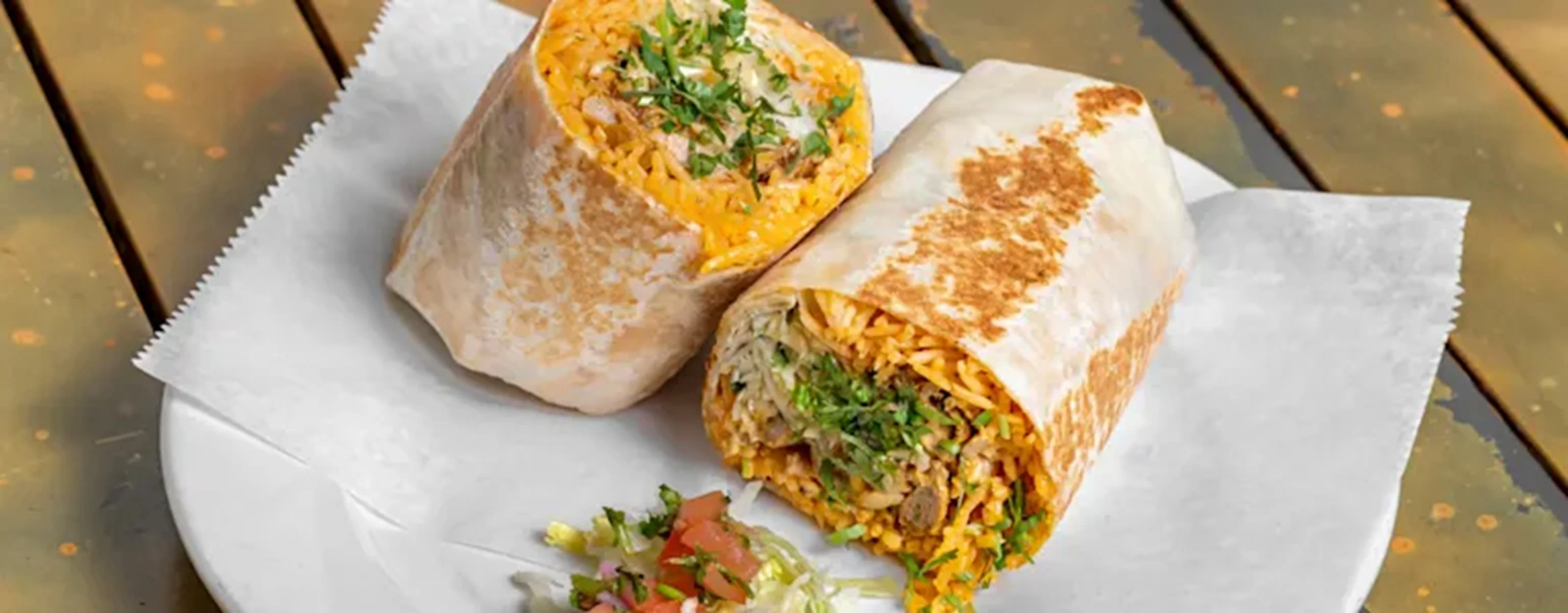 Two burritos are served on a white plate, featuring a mixture of rice, meat, and fresh herbs. A side of diced tomatoes and lettuce is also included.