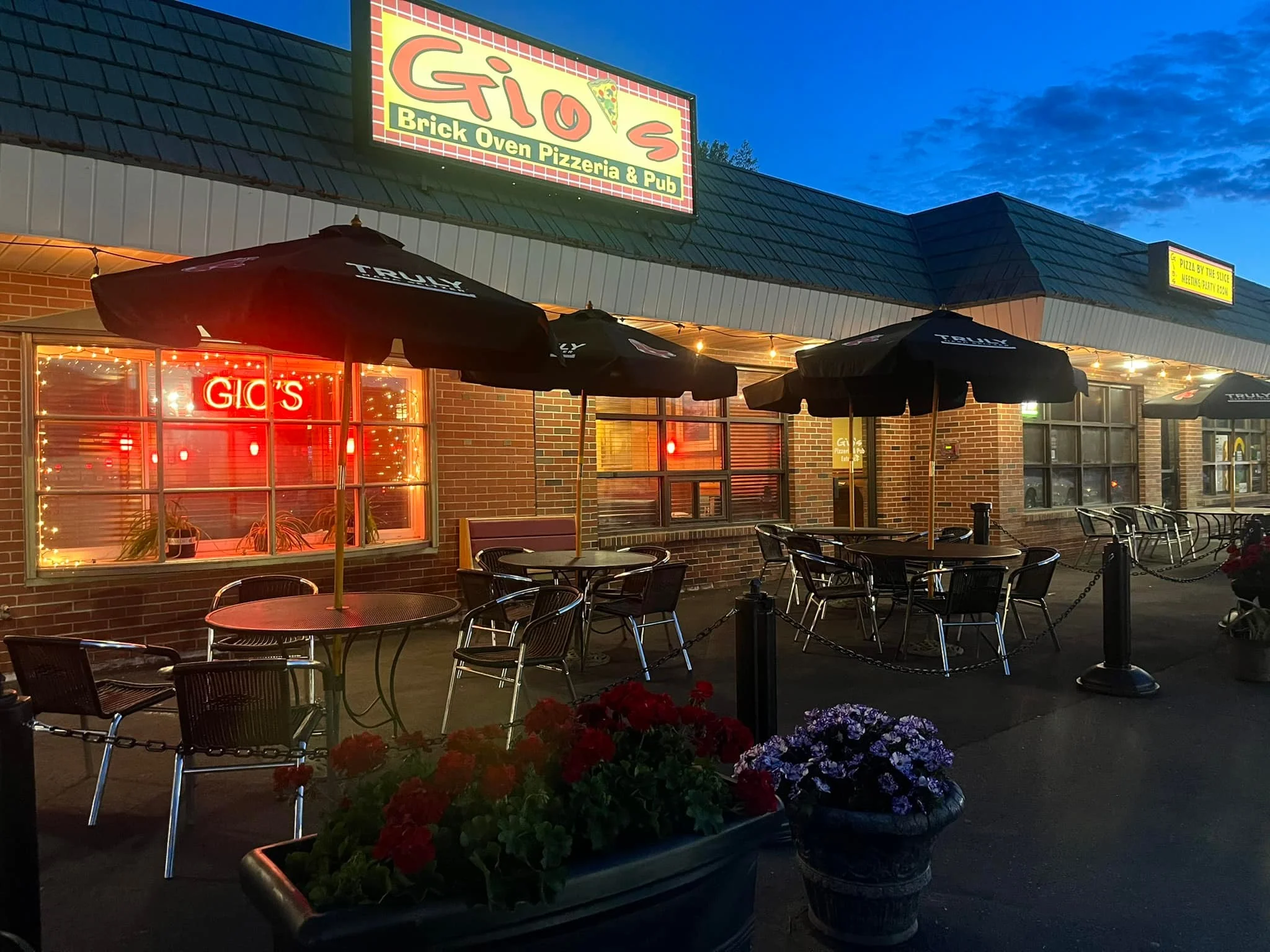 An outdoor dining area of a pizzeria featuring black umbrellas over tables, with colorful flower pots and a neon sign in the window.