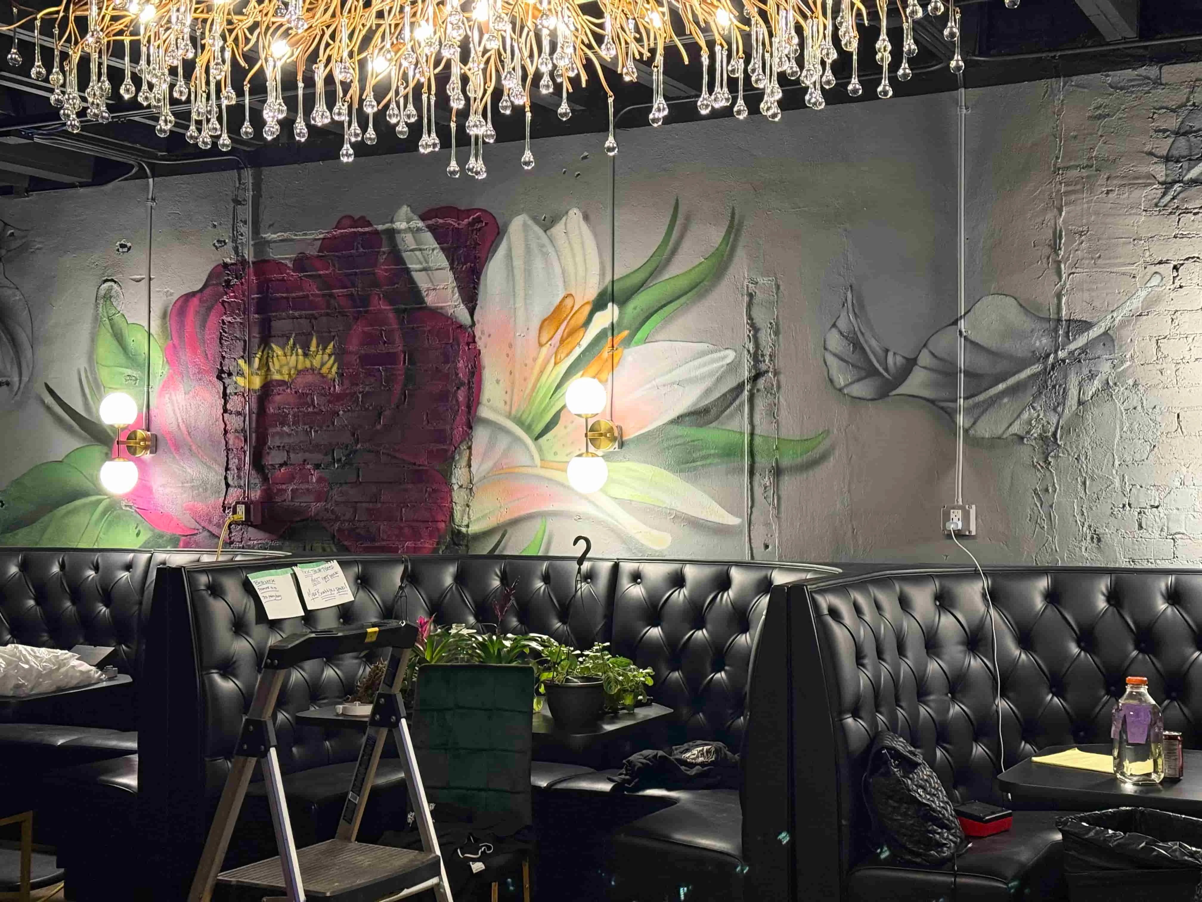 An inviting indoor seating area featuring plush black booths with a colorful floral mural in the background. Elegant lighting fixtures hang from the ceiling, enhancing the ambiance.
