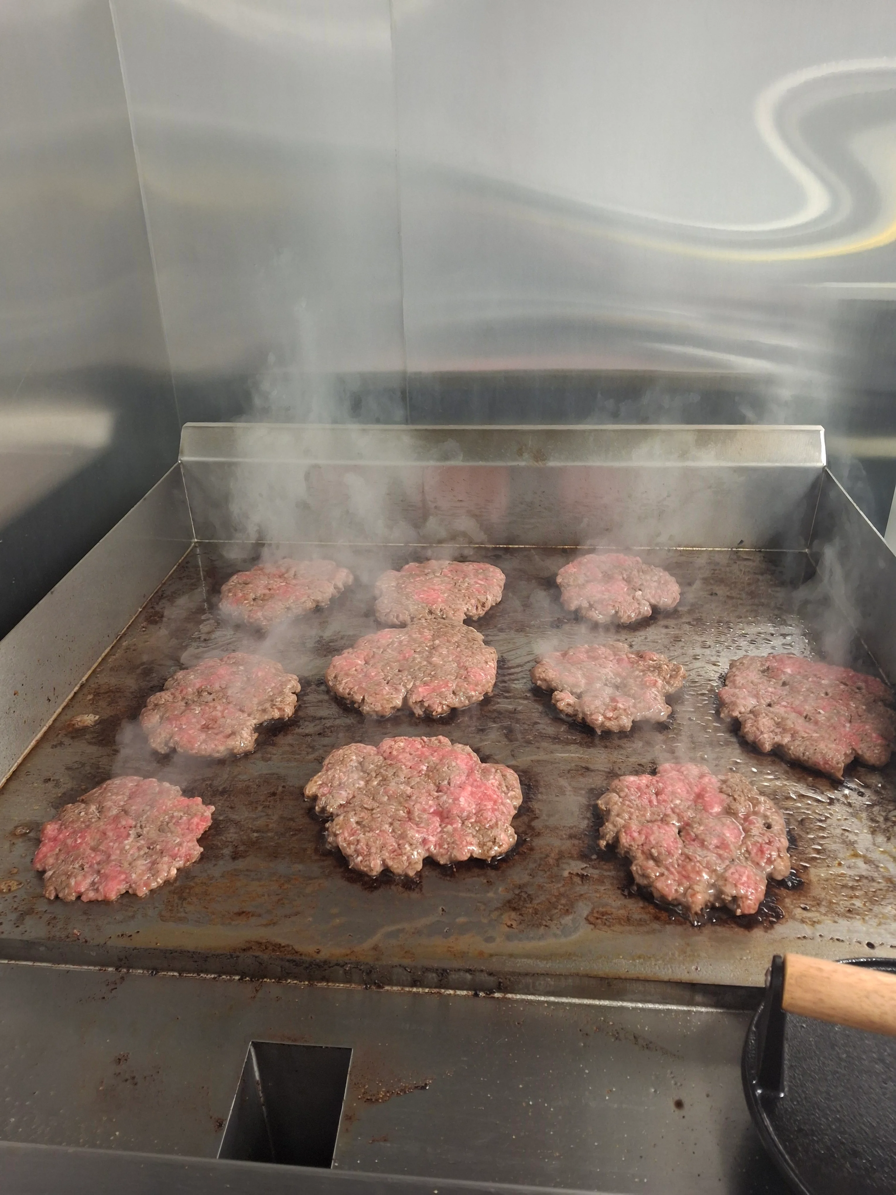 Multiple beef patties frying on a hot grill, with steam rising around them.