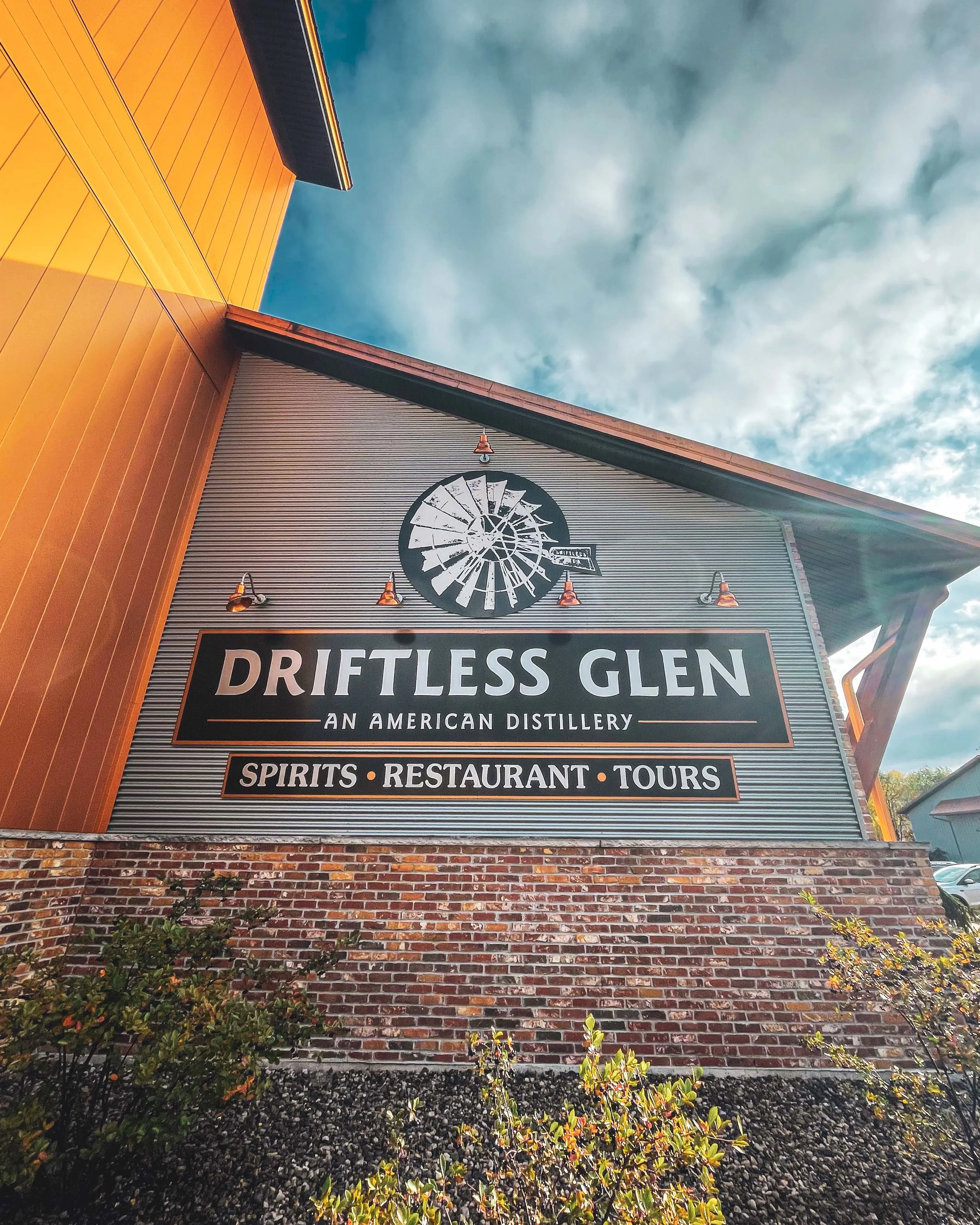The Driftless Glen entrance sign shines with sunlight and blooming bushes in front of it