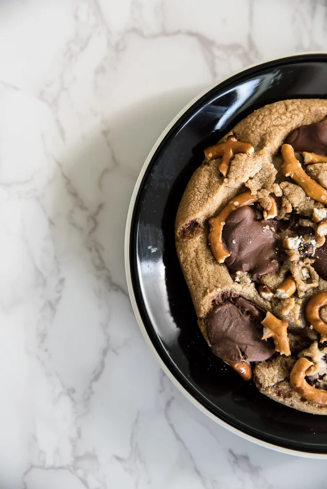 A large chocolate chip cookie topped with pretzel pieces sits on a black plate. The background is a white marble surface, enhancing a cozy, indulgent feel.