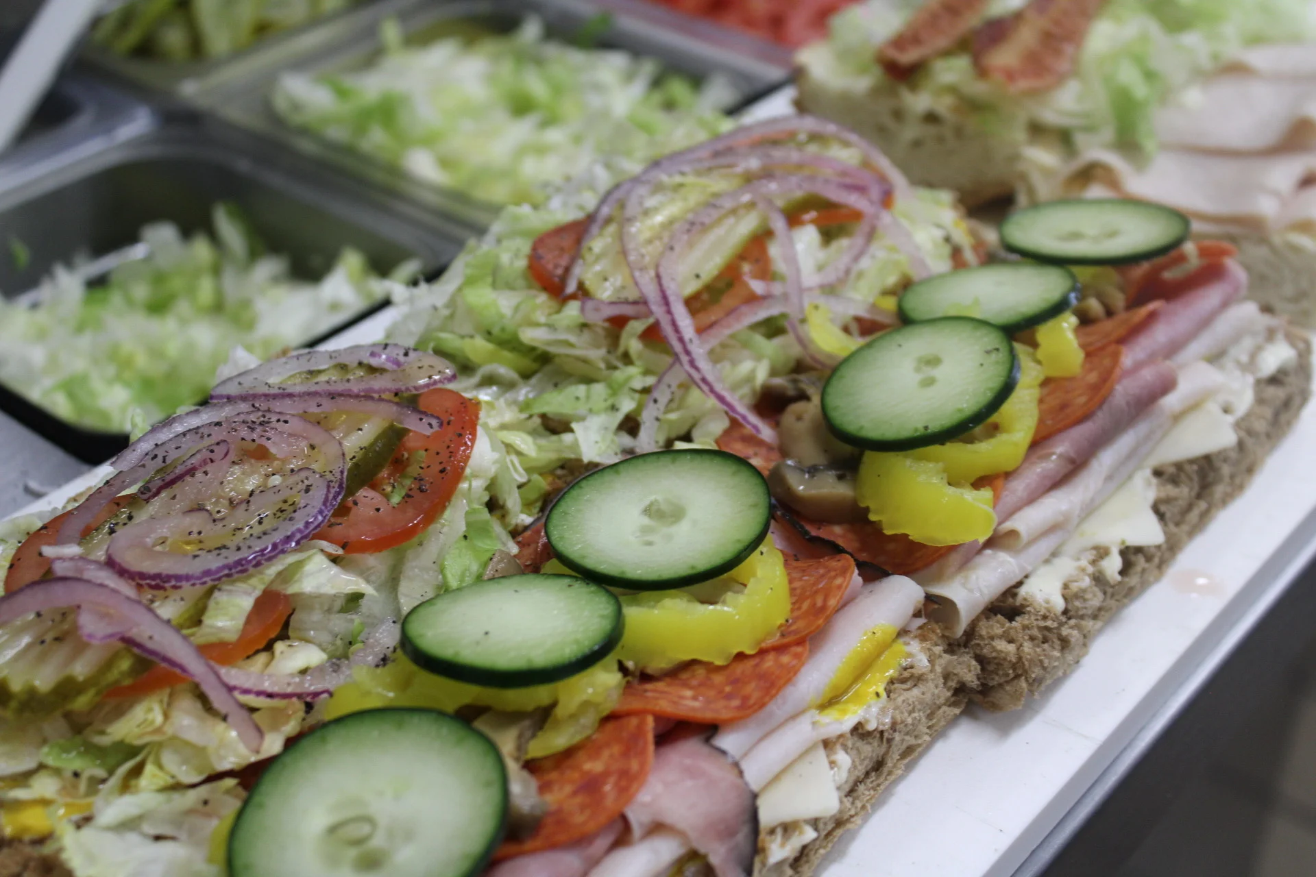 A large sandwich filled with layers of meats, fresh vegetables, and various toppings, displayed on a counter in a food preparation area.
