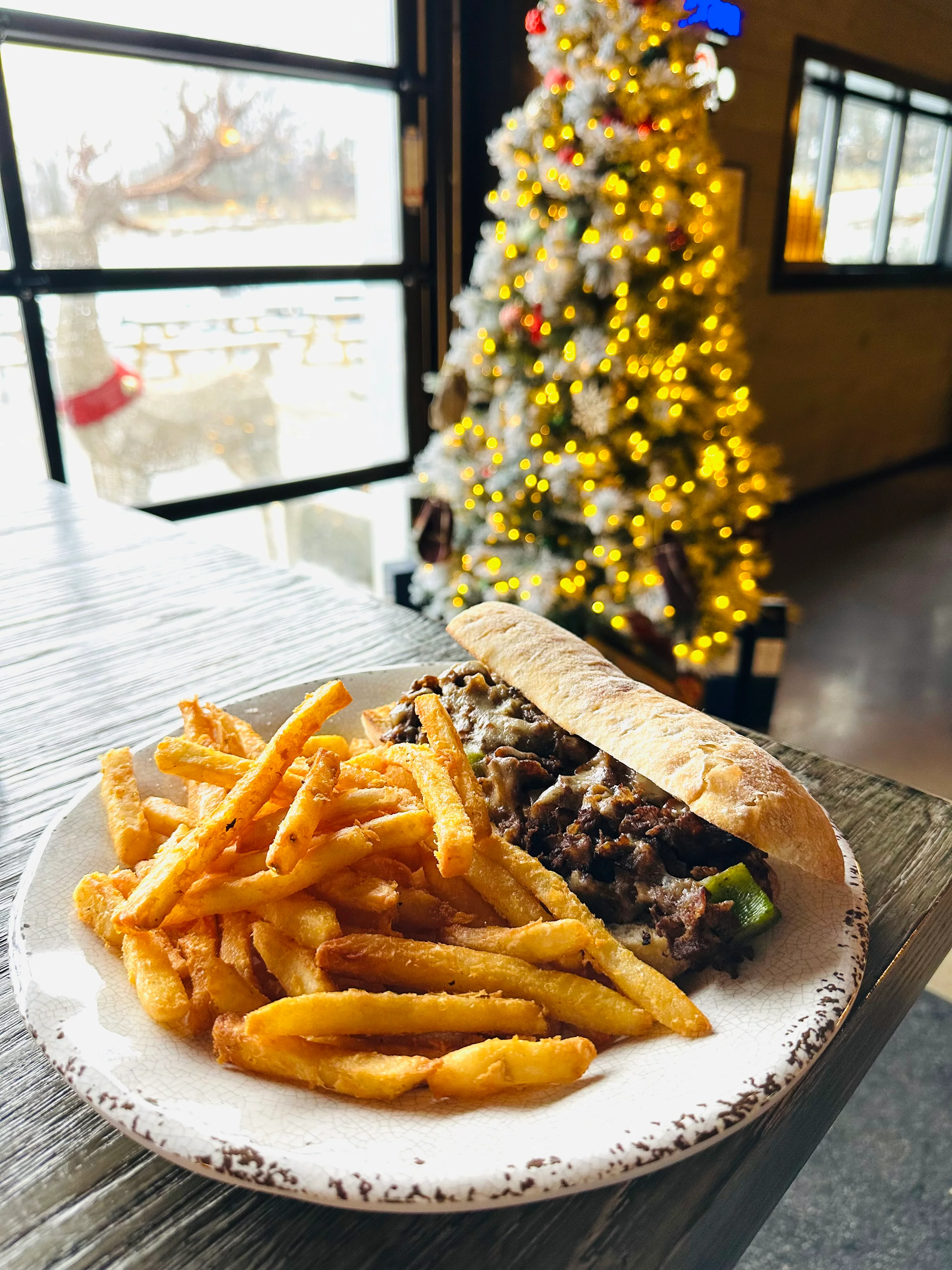 A plate featuring a sub sandwich filled with beef and cheese, accompanied by a side of golden fries. In the background, a decorated Christmas tree glows with lights.