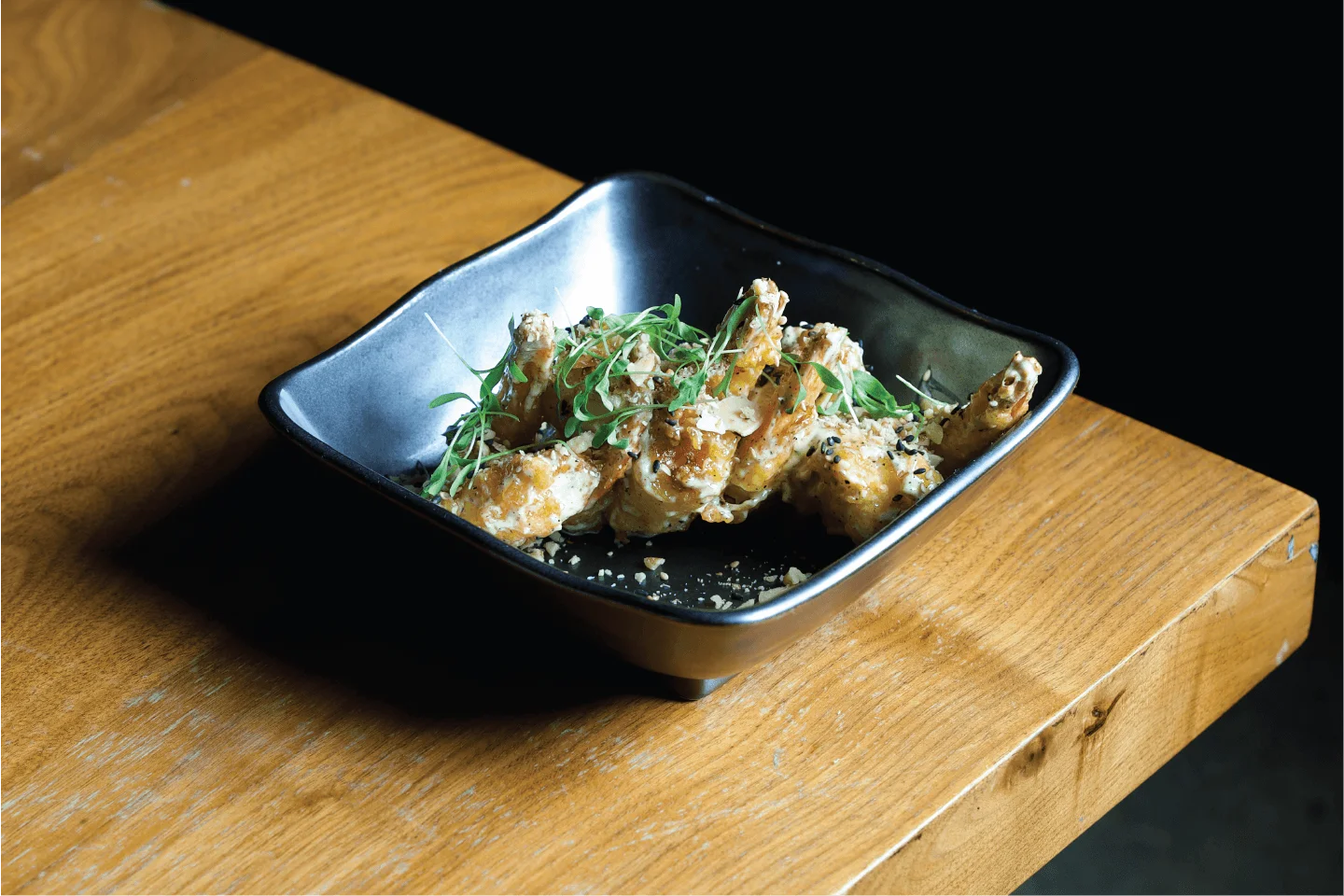 Crispy rock shrimp tempura tossed in a creamy sauce and topped with fresh microgreens and sesame seeds in a dark square bowl.