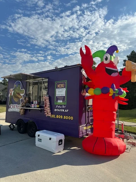 A colorful food truck featuring a cartoon alligator wearing a hat and holding a lobster. The truck is purple and advertises Cajun authentic foods with a catchy slogan.