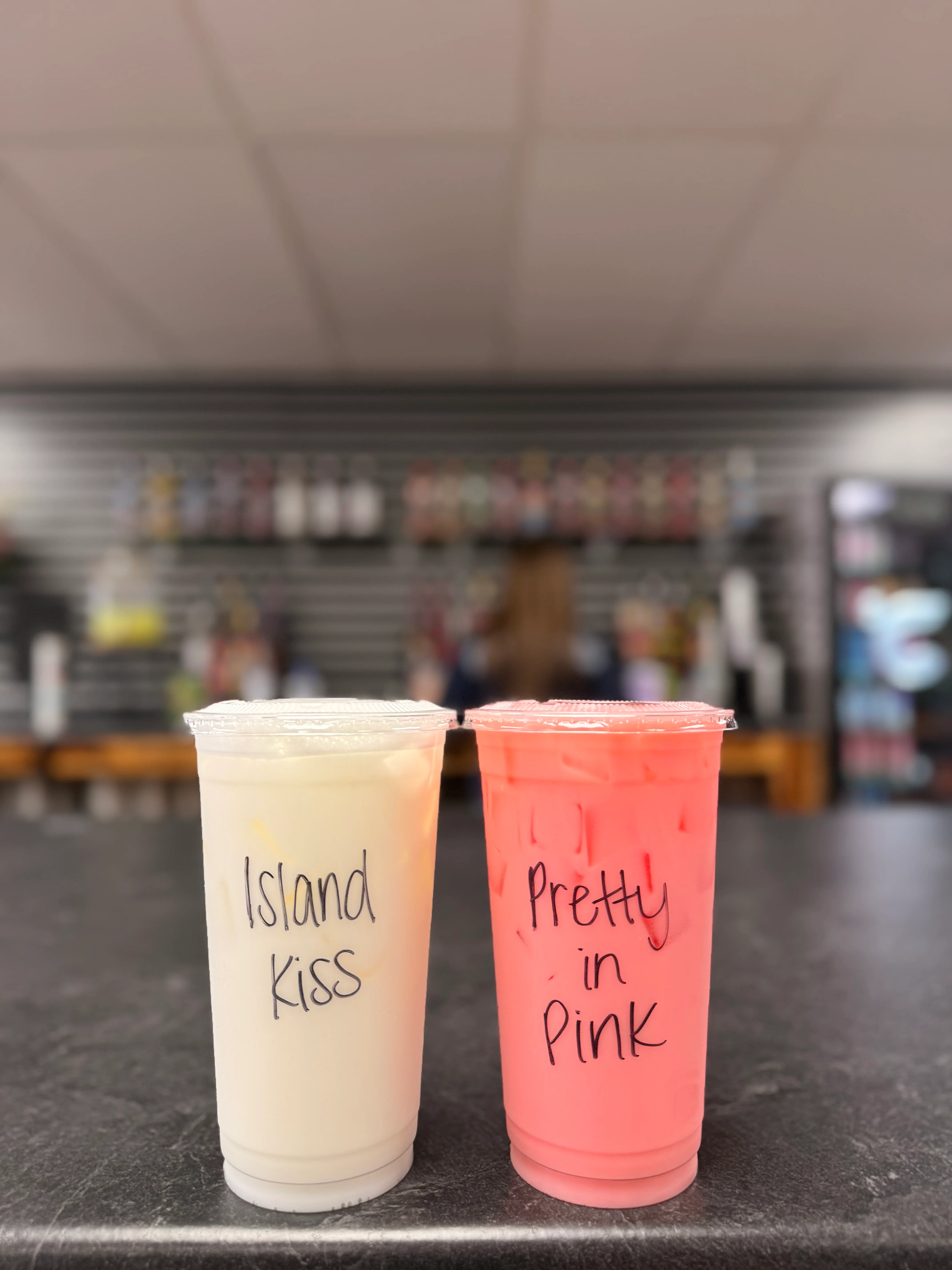 Two colorful beverage cups on a counter. The left cup is white with 'Island Kiss' written on it, and the right cup is pink with 'Pretty in Pink' labeled.