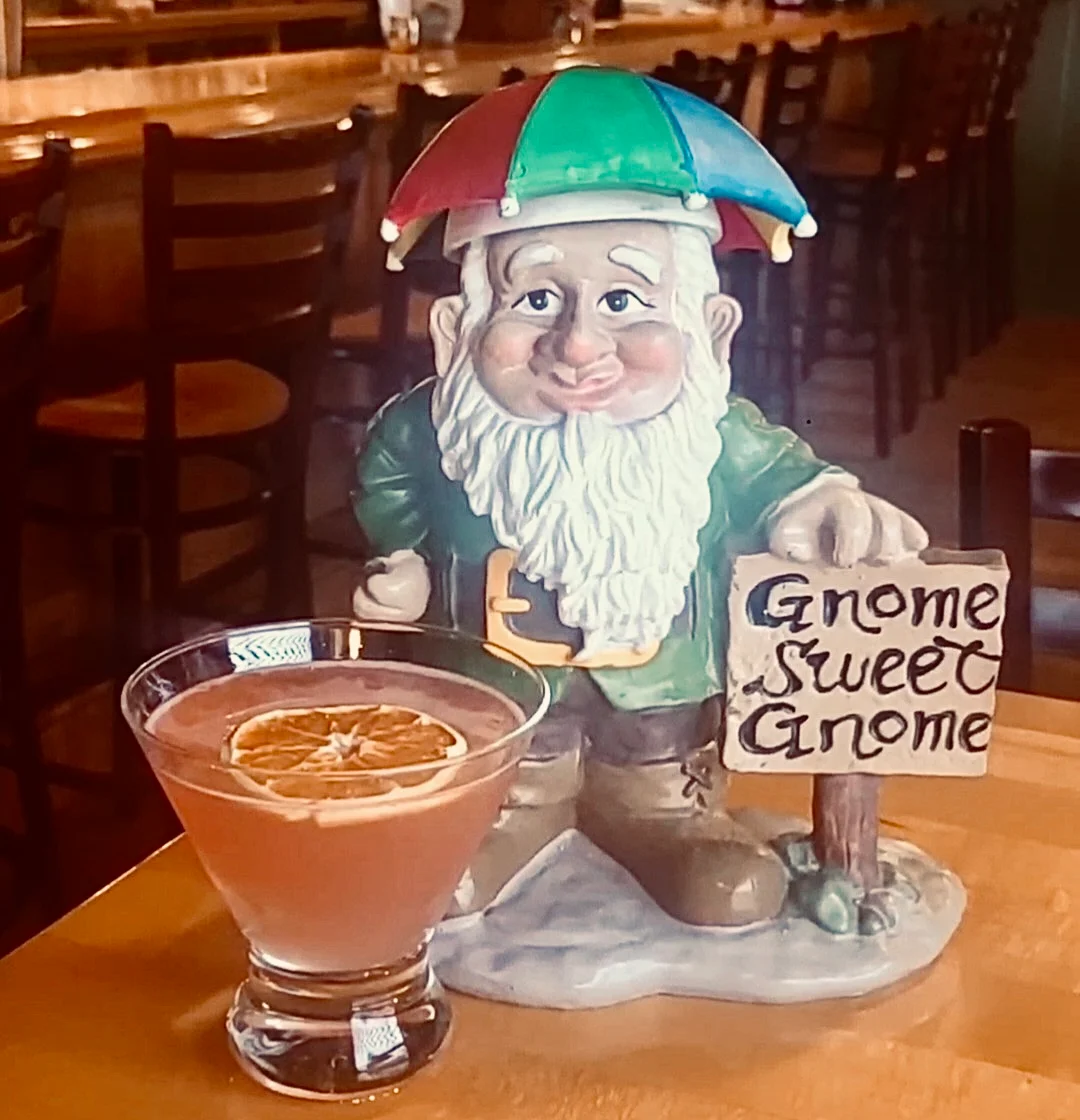 A cheerful gnome figurine holds a sign reading 'Gnome Sweet Gnome' next to a pink cocktail garnished with a dried orange slice. The setting showcases wooden bar stools in the background.
