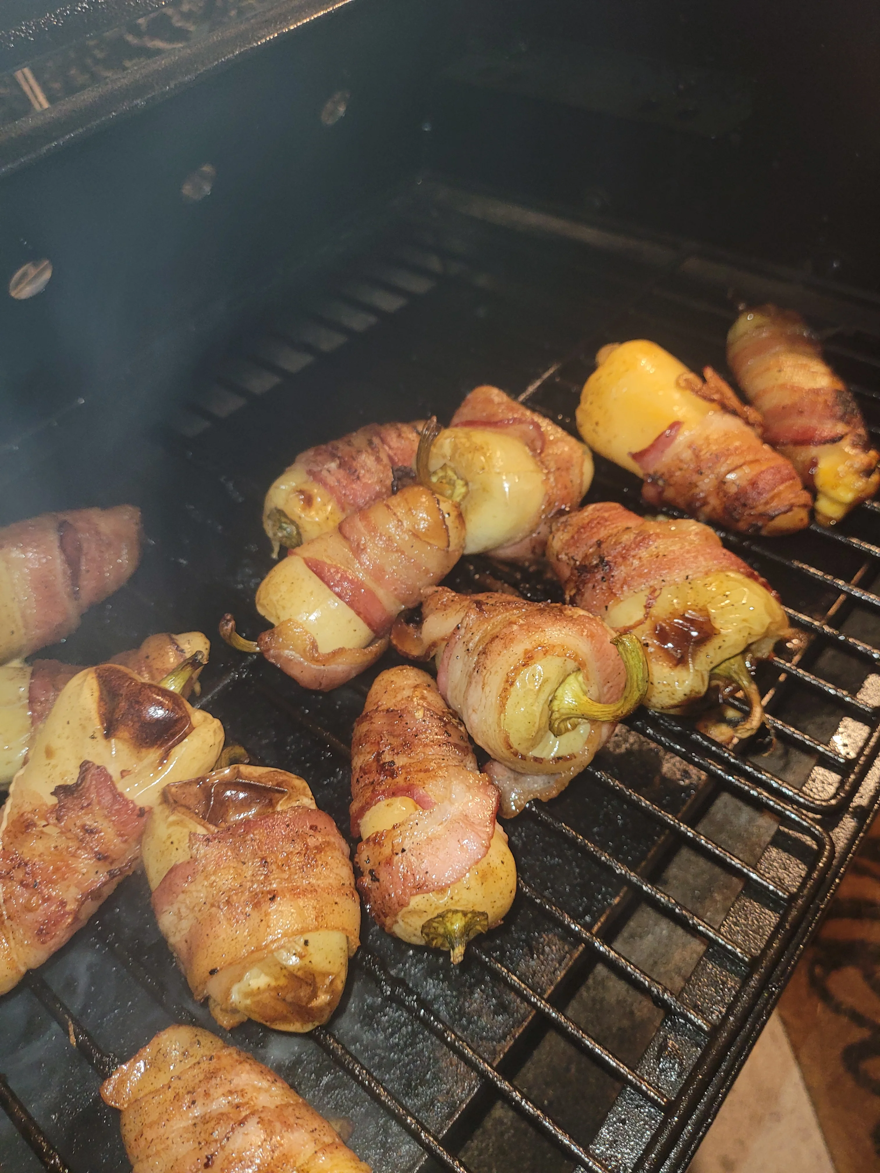 Bacon-wrapped jalapeño peppers are grilling, with some smoke rising from the grill.