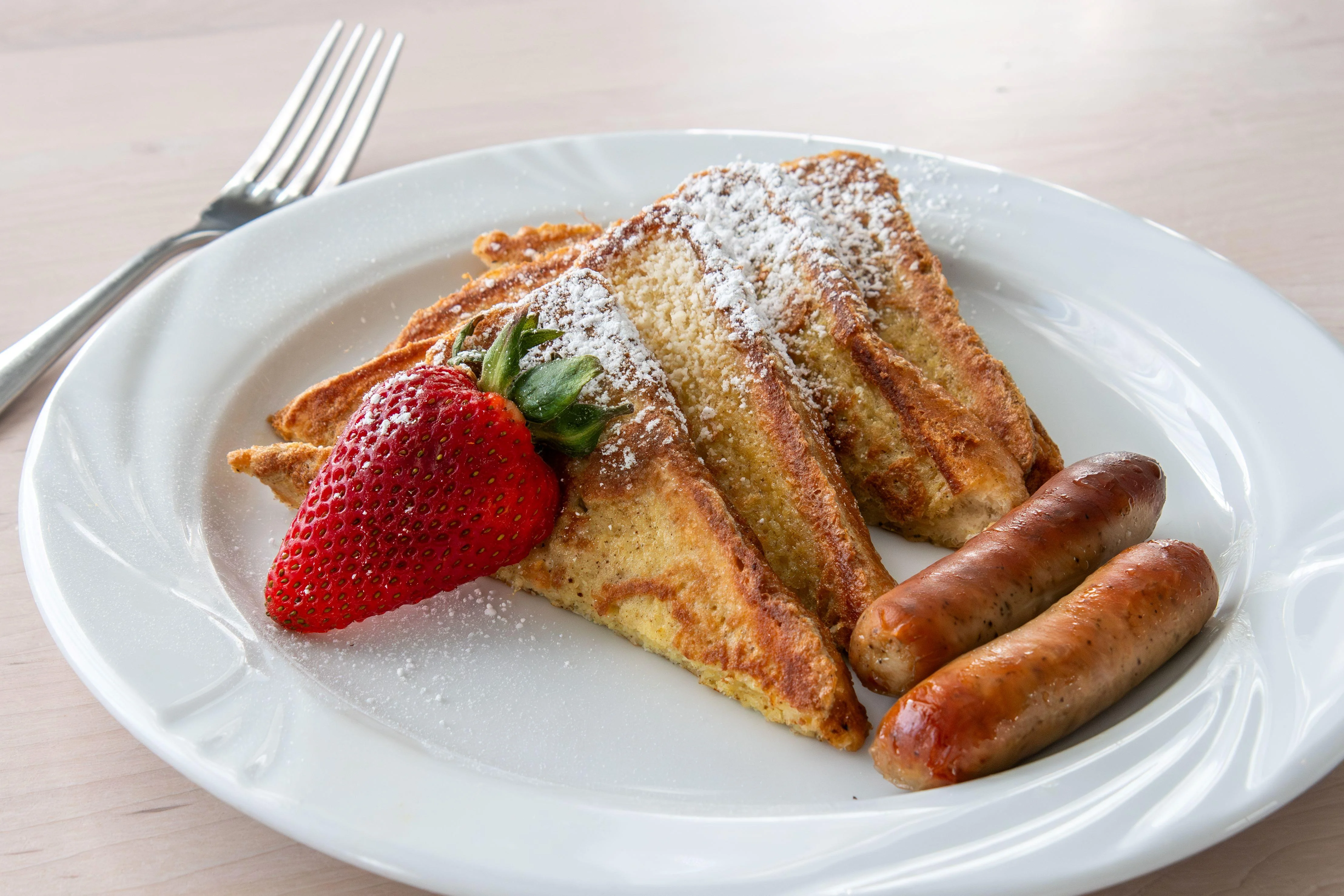 A plate featuring three slices of French toast dusted with powdered sugar, accompanied by a strawberry and two sausage links.