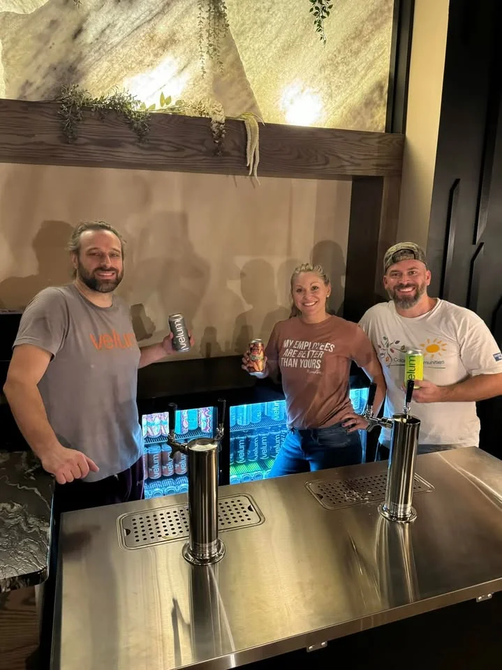 A casual gathering at a bar with three people standing in front of a drink dispenser, each holding a beverage. The setting has a modern and friendly vibe.