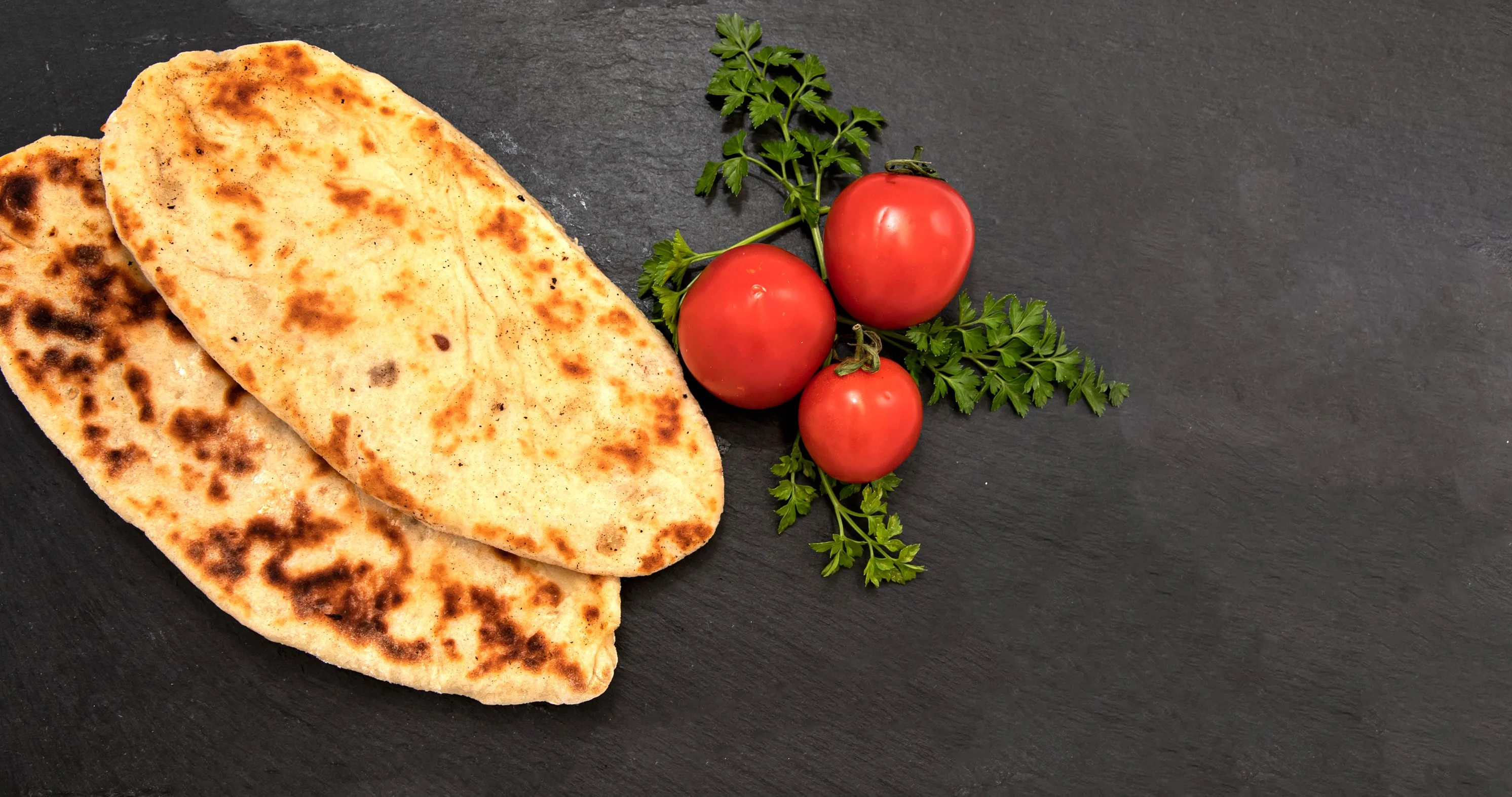Two pieces of freshly baked naan bread placed next to ripe tomatoes and parsley on a slate background.