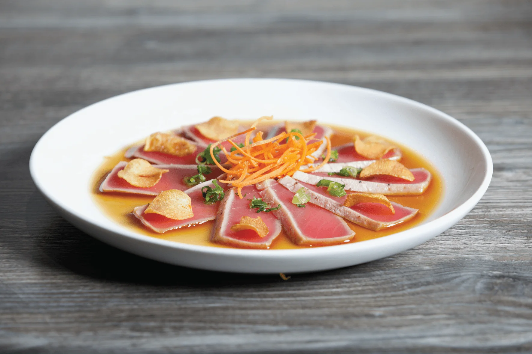 Seared tuna tataki slices arranged in a circle with garlic chips, scallions, and shredded carrots in a light citrus sauce.