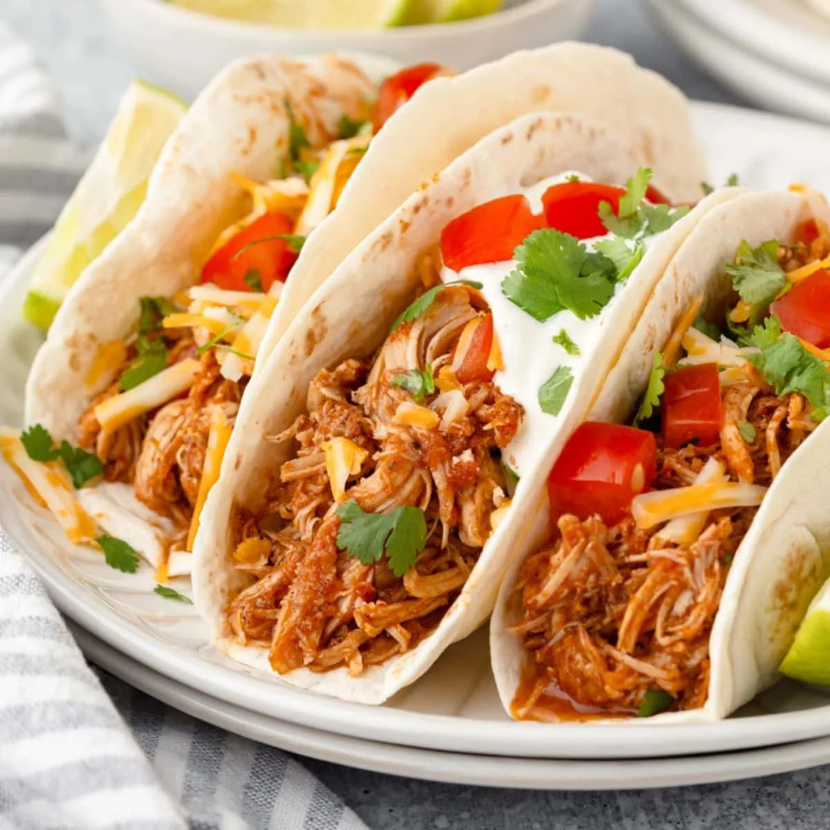 Three tacos filled with shredded meat, topped with diced tomatoes, shredded cheese, sour cream, and fresh cilantro. Served with lime wedges.