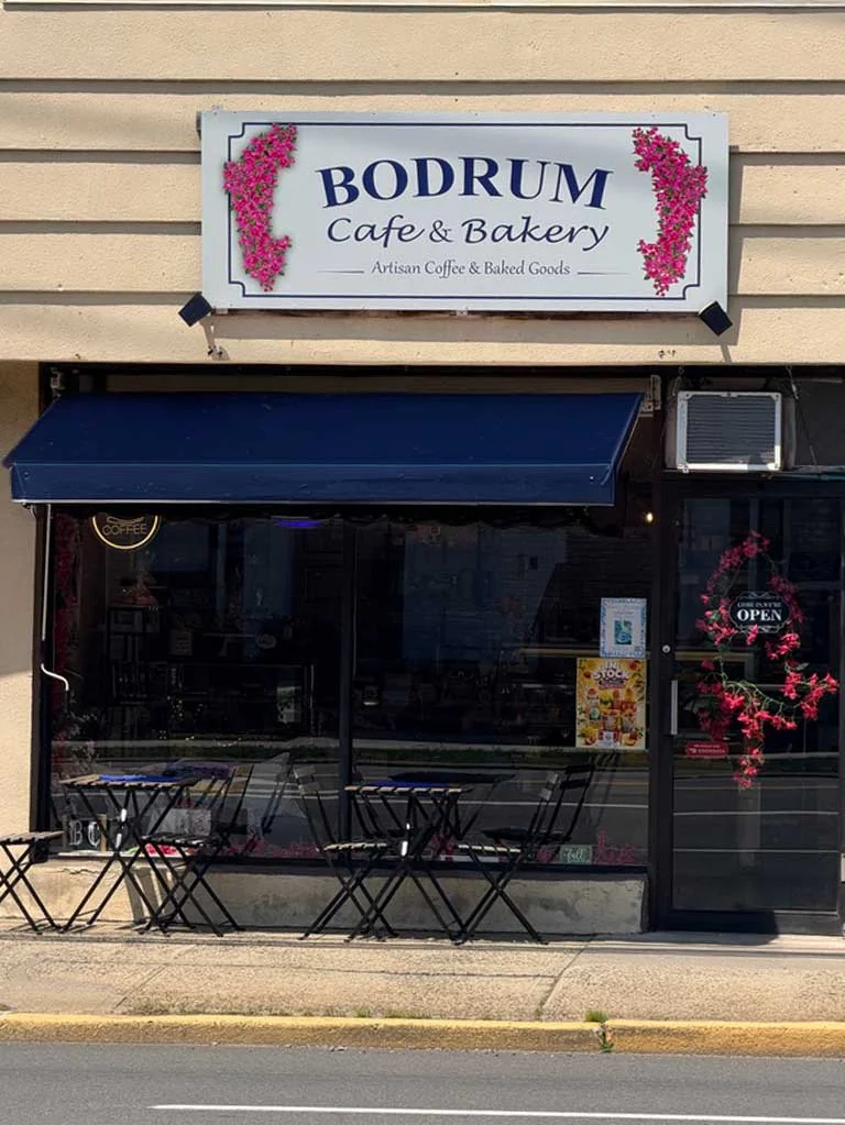 A colorful logo for 'Bodrum Cafe & Bakery' featuring a scenic view of a coastline with a castle and a sailboat, framed by vibrant pink flowers.