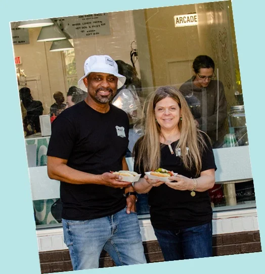 Two women holding a tray of hot dogs with toppings, smiling together.