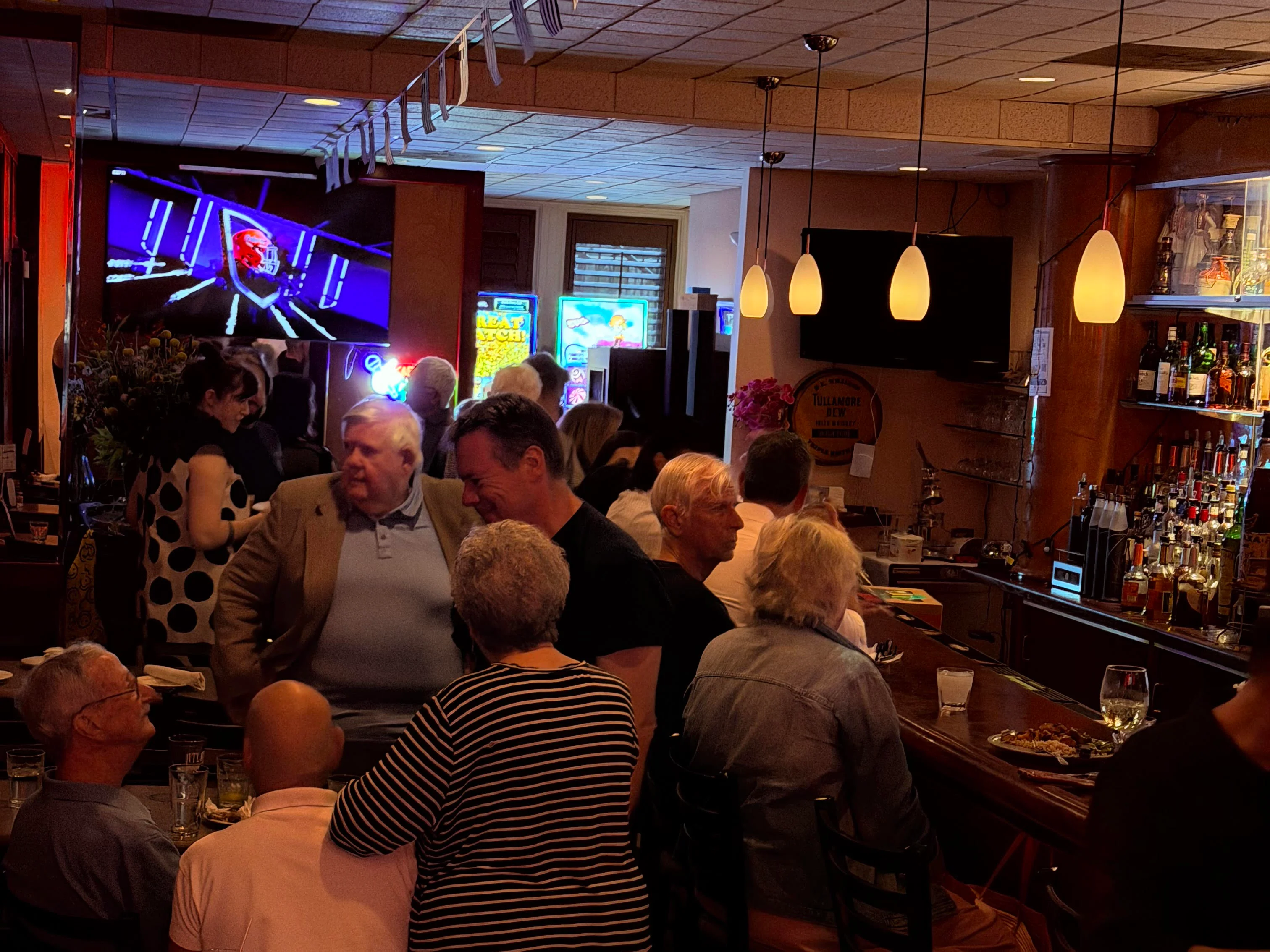 Busy restaurant dining room with patrons, pendant lights, bar, and television