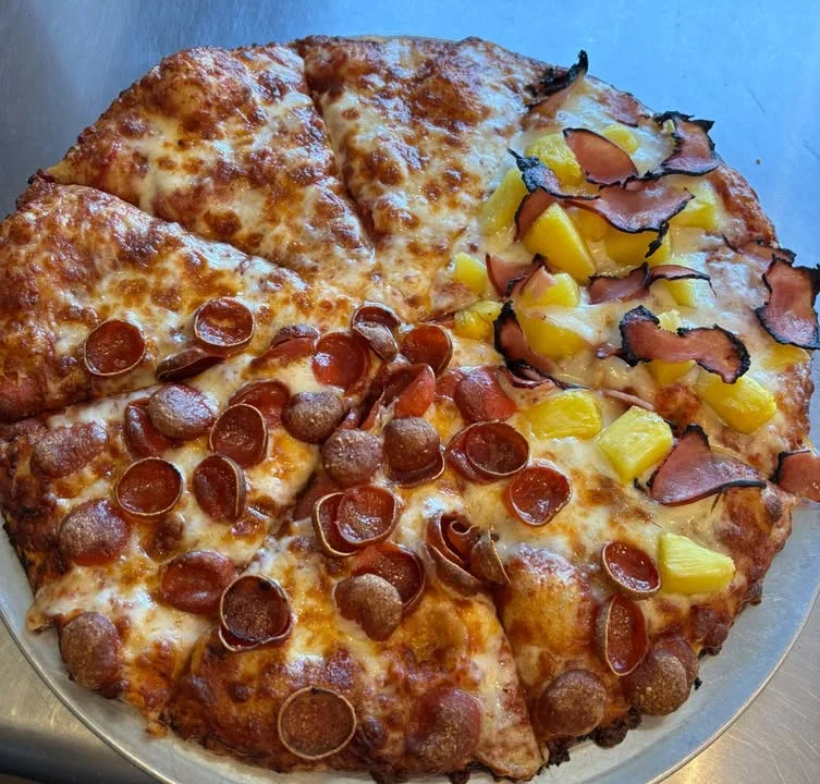 A half-and-half pizza featuring pepperoni and sausage on one side, and ham and pineapple on the other, with melted cheese throughout.