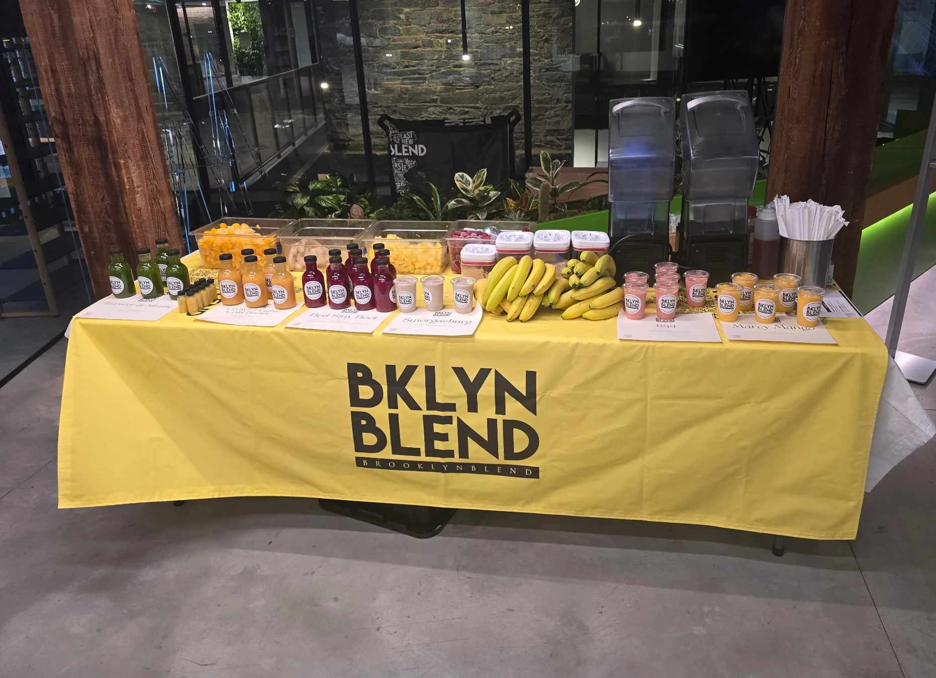 A yellow tablecloth with "BKLYN BLEND" branding covers a table displaying fresh juices, bananas, and cut fruit containers, creating a vibrant, healthy atmosphere.