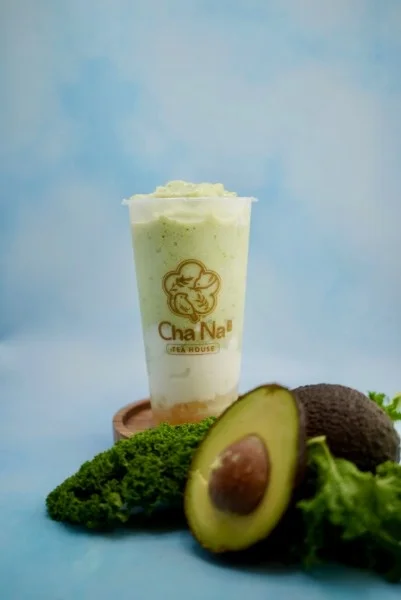 A creamy green beverage topped with whipped foam, served in a branded cup. Fresh avocados and leafy greens are placed beside it.