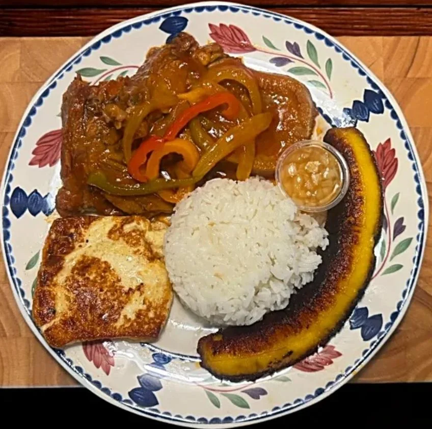 A hearty plate of chicken in sauce with rice, fried cheese, and a slice of fried plantain, served with a small cup of sauce.