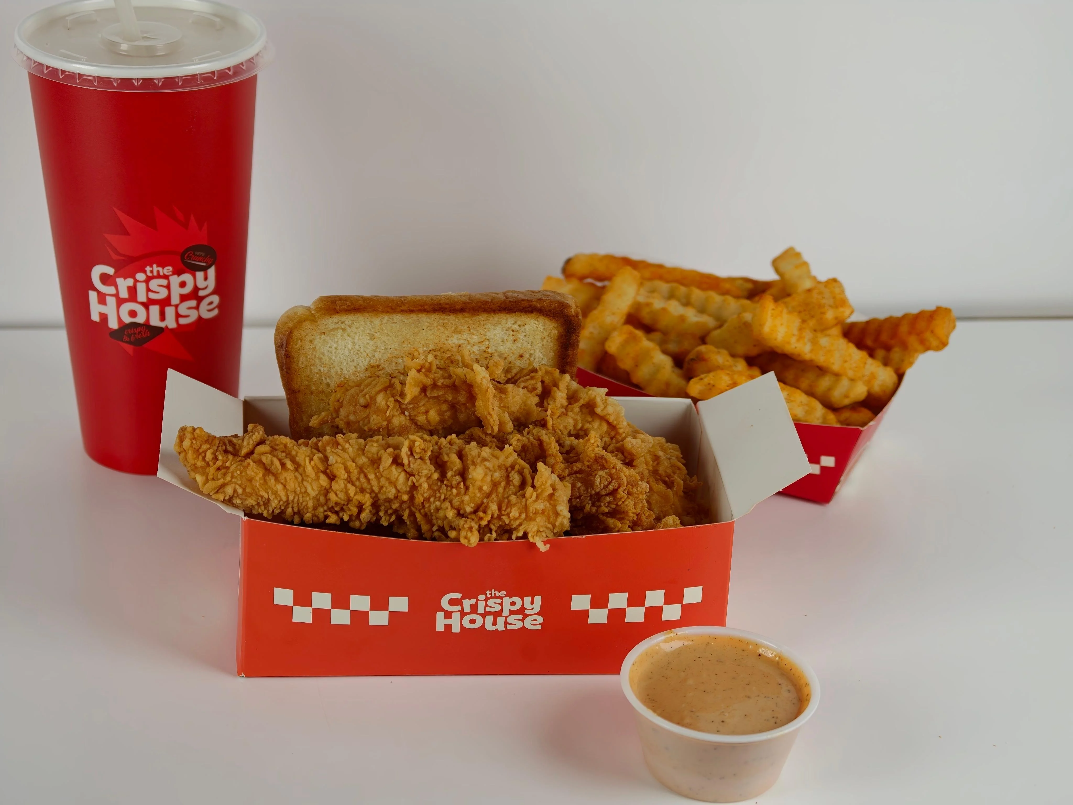 A meal featuring crispy fried chicken strips, a slice of white bread, crinkle-cut fries, and a small cup of dipping sauce. Accompanied by a large drink.
