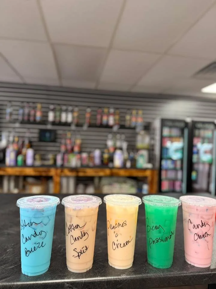Five colorful drinks in clear cups, labeled with different flavors, sitting on a counter. The drinks are various pastel shades and feature toppings.