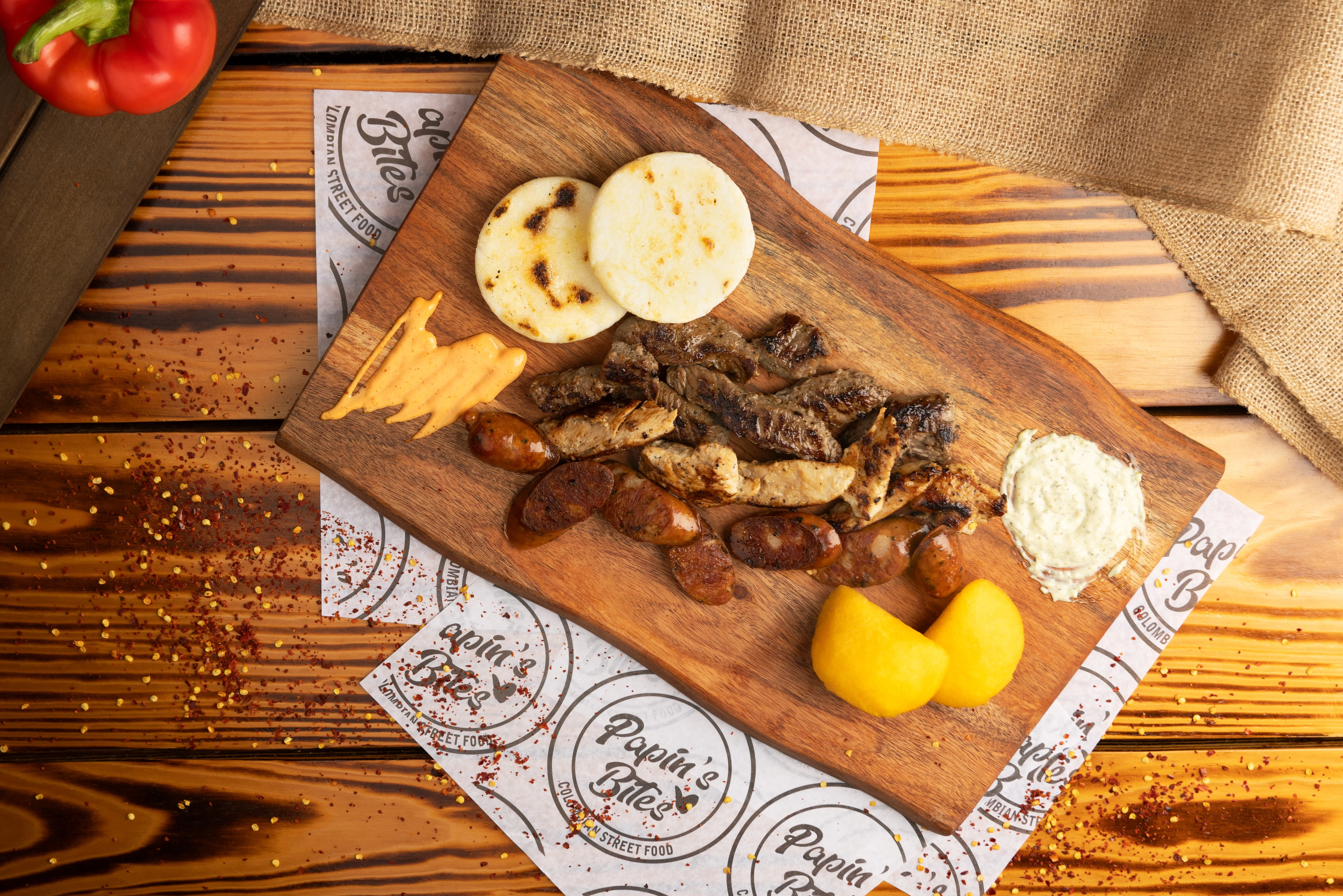 A wooden platter displaying grilled meats, two arepas, and sides including sauce and fried plantains, all served on a rustic table.