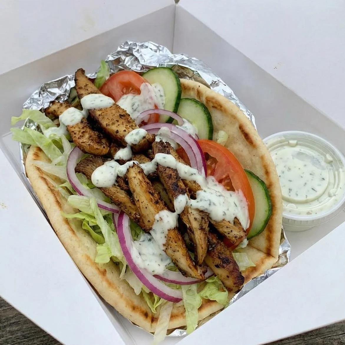 A box containing a flatbread filled with grilled chicken, lettuce, tomatoes, cucumbers, and onions, topped with a creamy dressing.