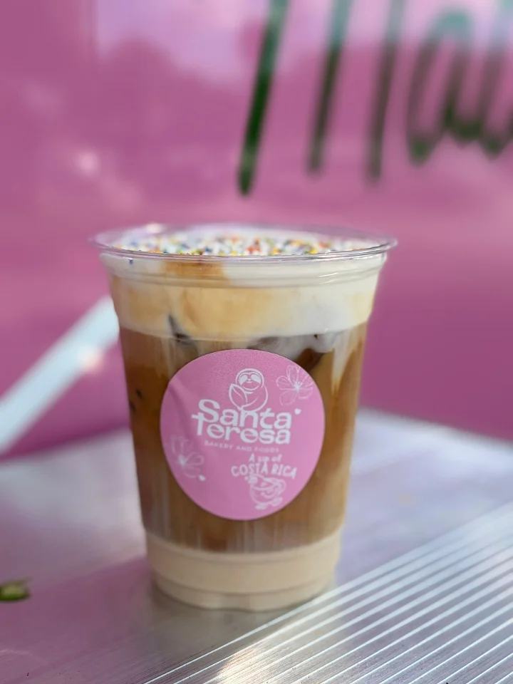 A decorated iced coffee topped with whipped cream and colorful sprinkles, presented in a clear cup with a pink logo.