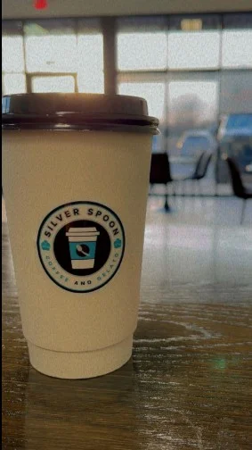 A paper coffee cup with the 'Silver Spoon' logo on a table in a cafe setting.