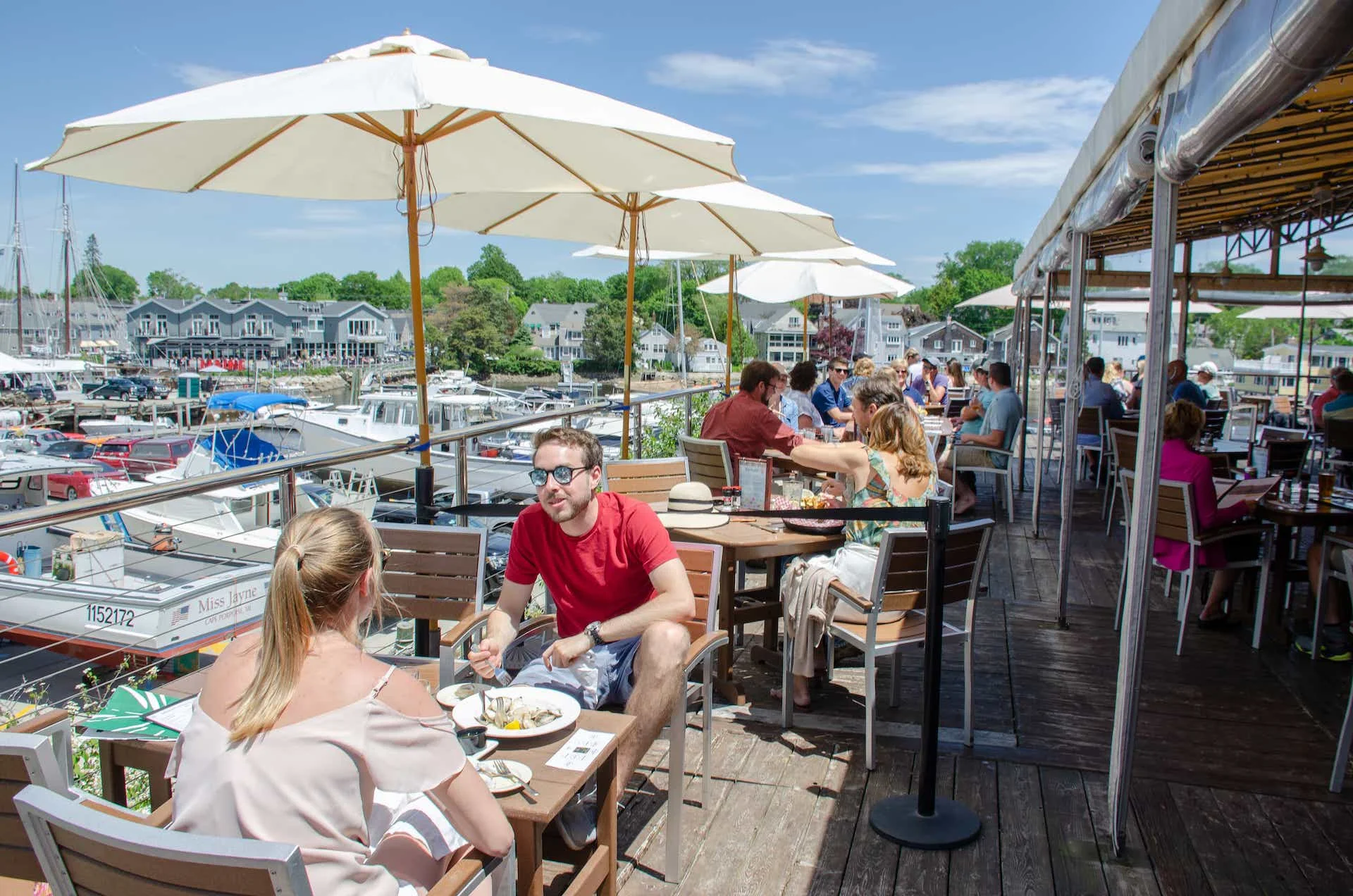 A lively waterfront restaurant with people dining at tables, enjoying the view of boats and the marina.