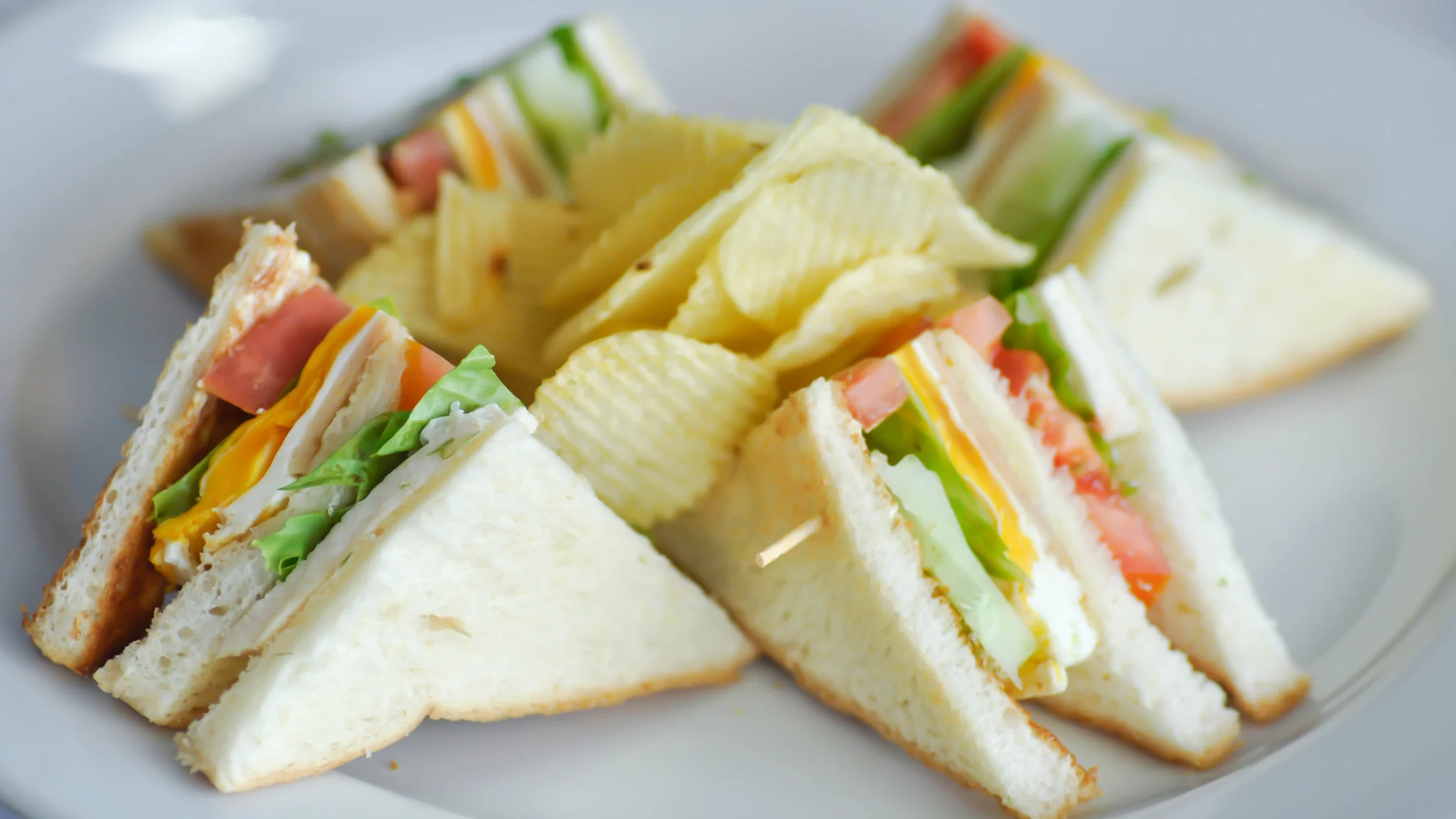 Assorted club sandwiches made with soft white bread, layered with lettuce, tomato, cheddar cheese, turkey, and ham, arranged in wedges around a pile of ridged potato chips on a white plate.