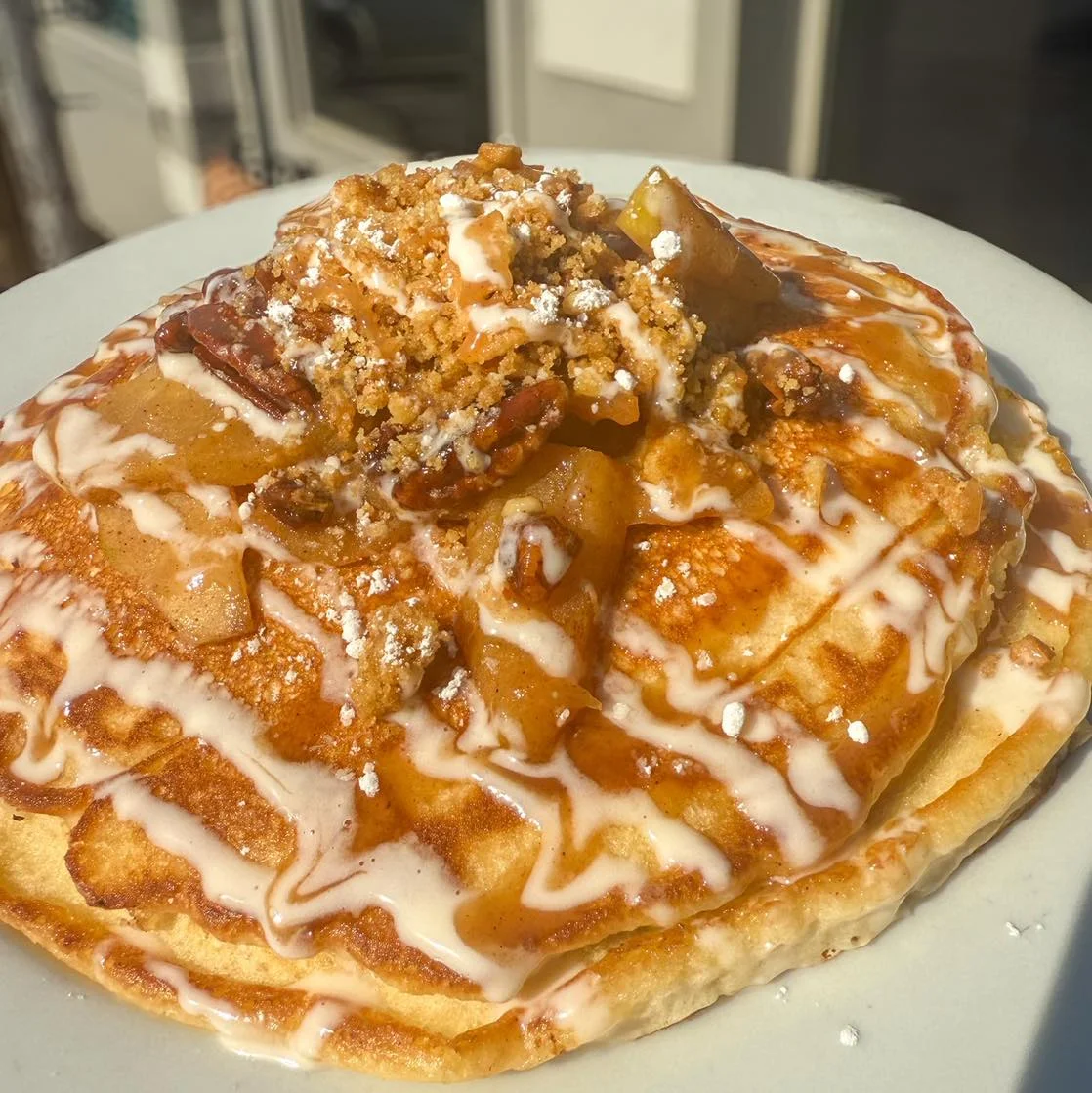 A stack of pancakes topped with caramelized apples, crumbly topping, and a drizzle of icing.