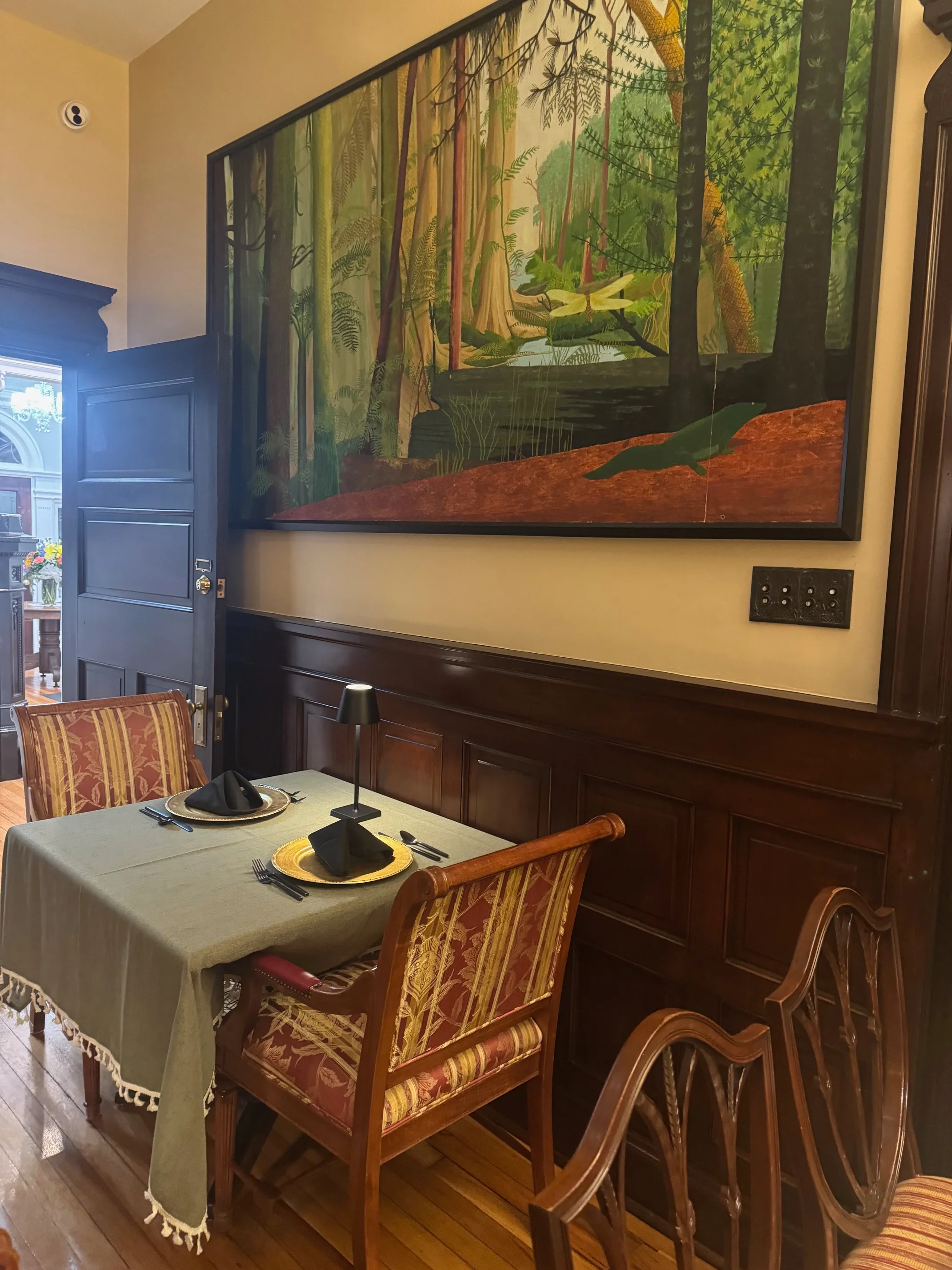 A dining setup featuring a table with neatly arranged plates and cutlery, accompanied by a vibrant landscape painting on the wall, creating a serene dining experience.