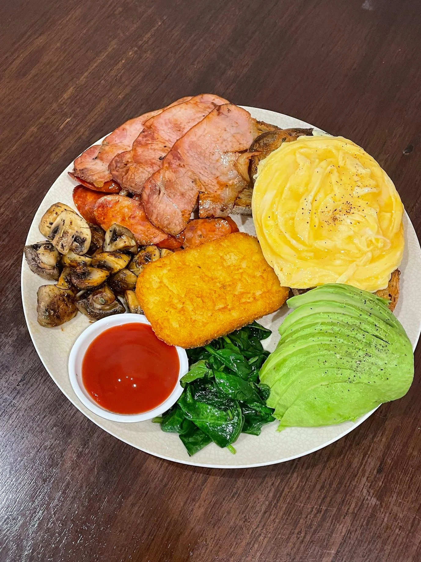 A hearty breakfast plate with slices of ham, sausage, hash brown, avocado, sautéed spinach, and mushrooms, served with a side of ketchup.