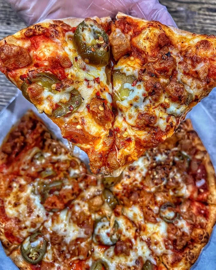 A hand holding a slice of pizza topped with jalapeños and meat, above another pizza showing similar toppings.