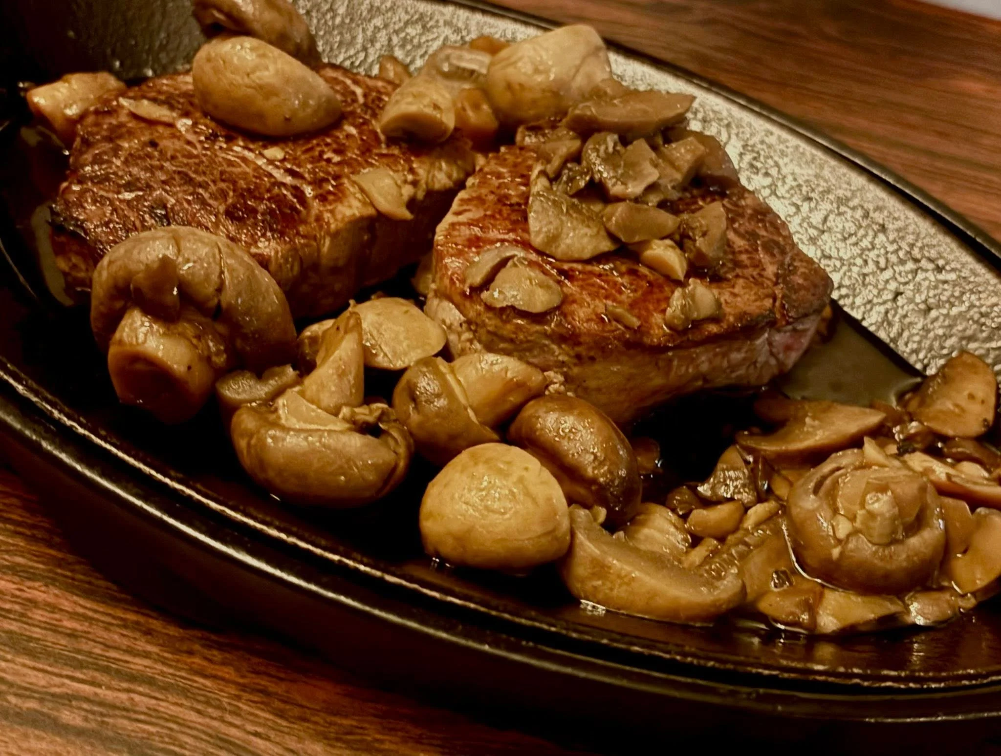 A sizzling steak dish topped with sautéed mushrooms on a black serving plate.