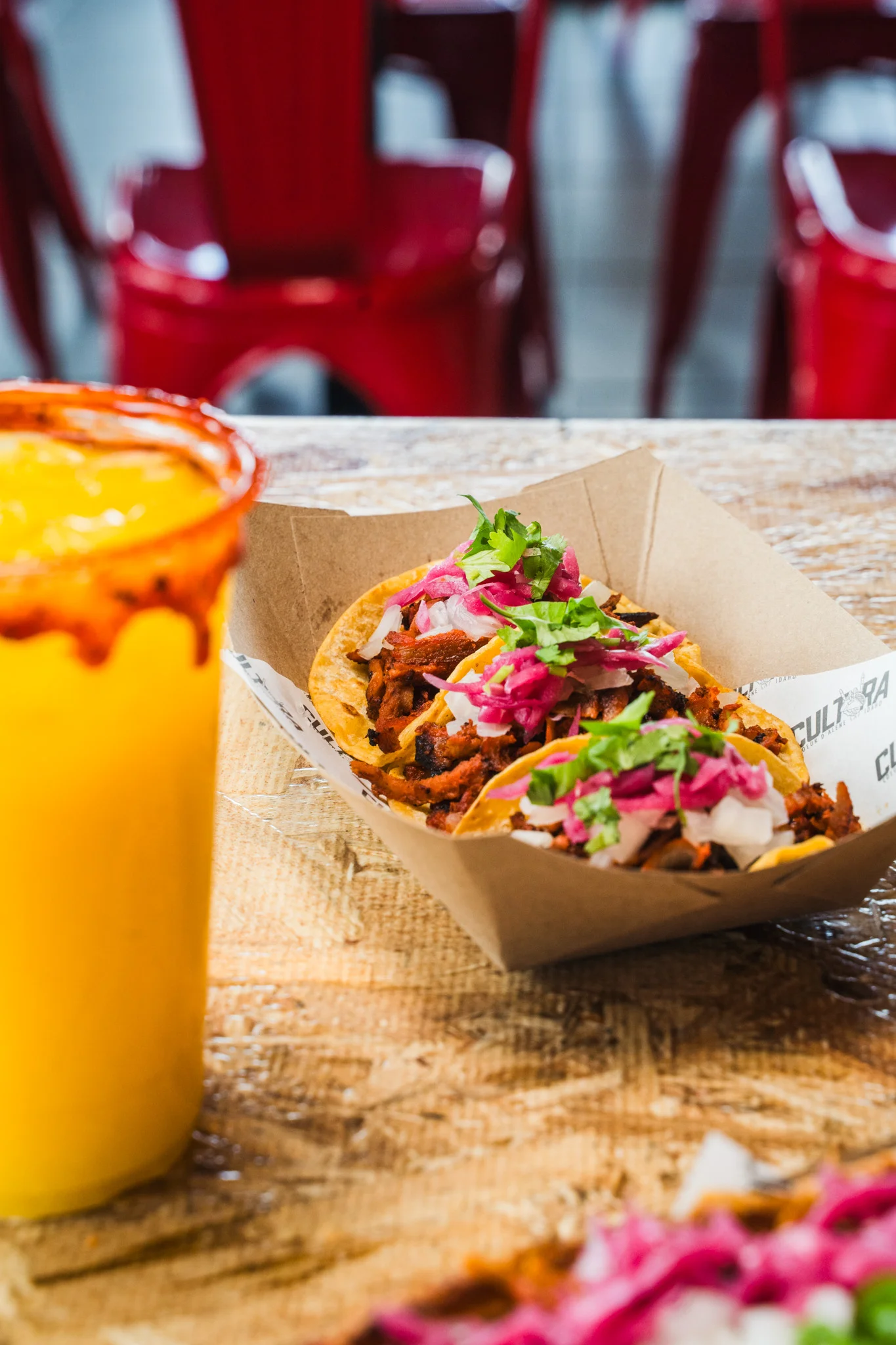 Three tacos topped with onions, cilantro, and pink pickled onions next to a drink.