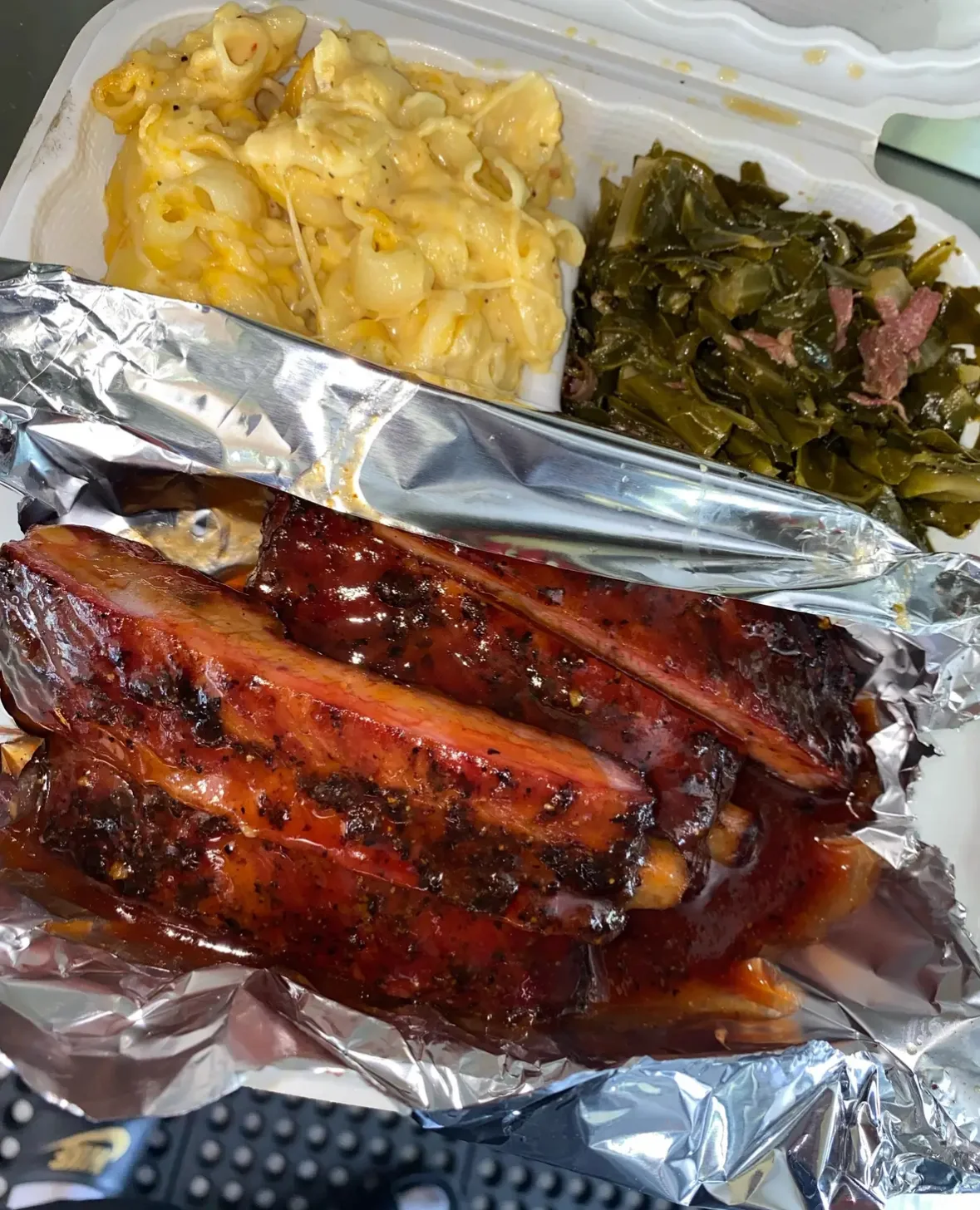 A serving of grilled meat ribs coated in barbecue sauce, accompanied by mac and cheese and collard greens in a takeout container.
