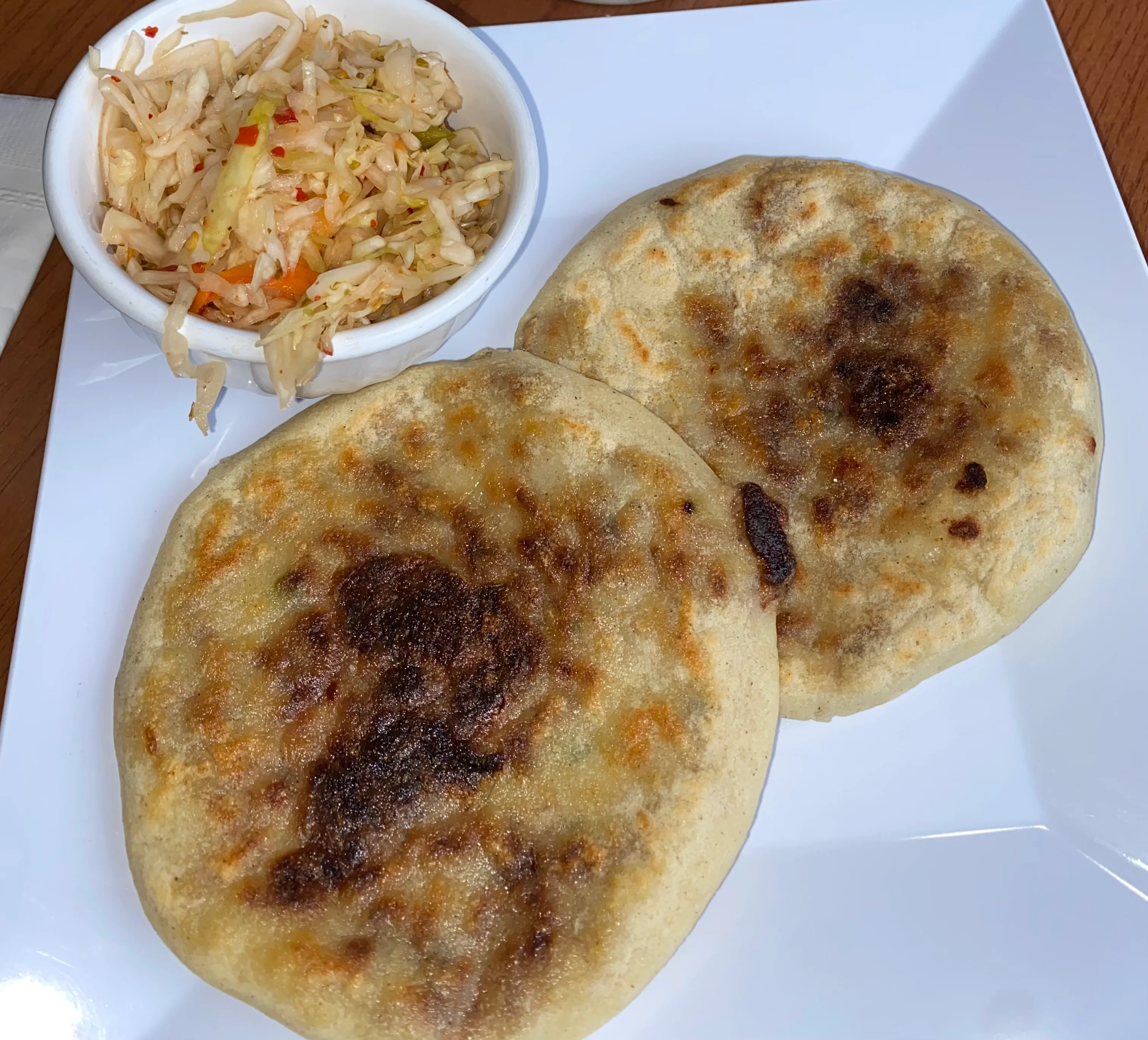 Two golden-brown pupusas served on a white plate, alongside a small bowl of coleslaw.