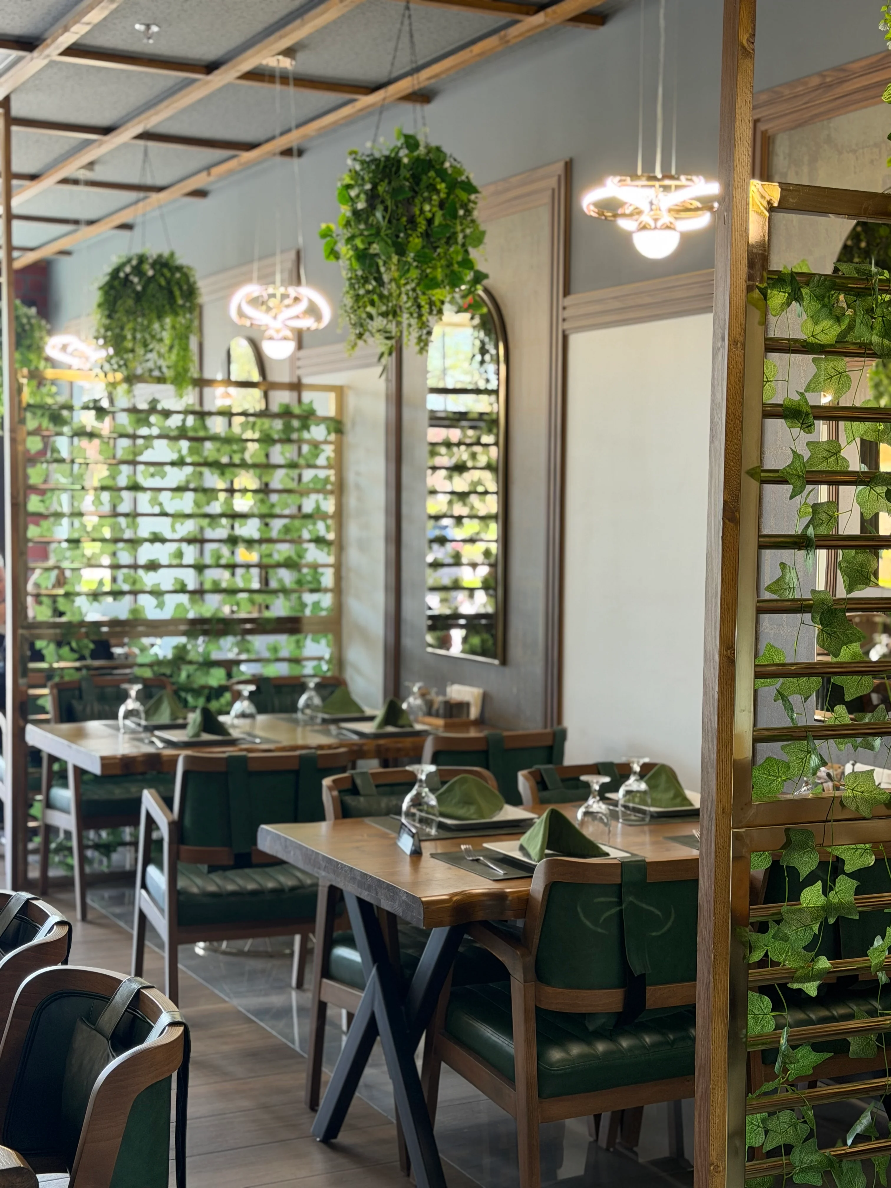 An elegant dining setting with green chairs, decorative plants, and stylish lighting fixtures. The tables are set with napkins and glassware.