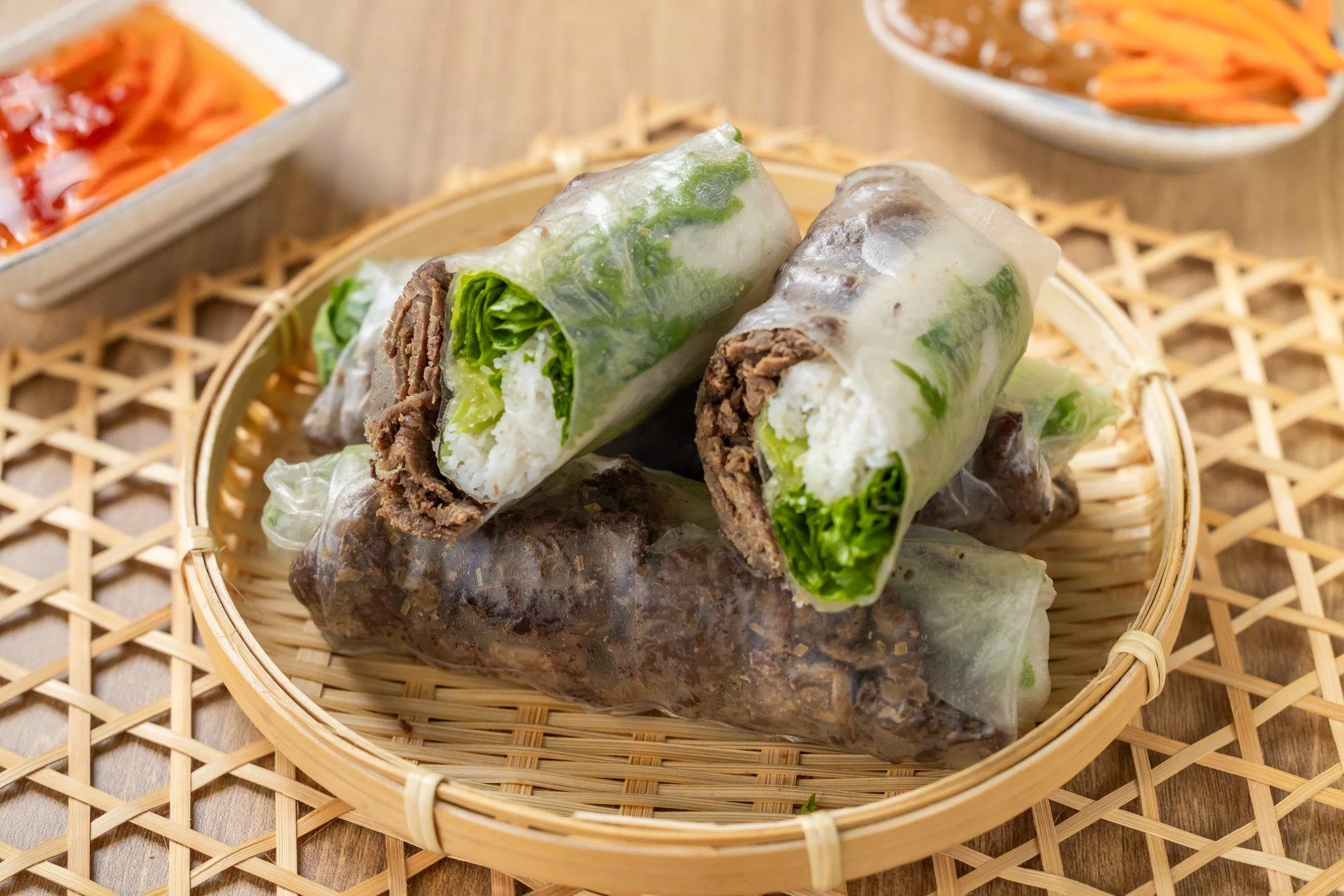 A close-up of rolled rice paper wraps with layers of lettuce and beef. The basket holds additional sides of sauce and pickled carrots.