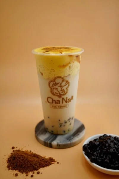 A refreshing drink featuring a creamy layer with a scoop of ice cream on top. It is served in a clear cup with a logo, alongside a small bowl of tapioca pearls and a heap of brown sugar.