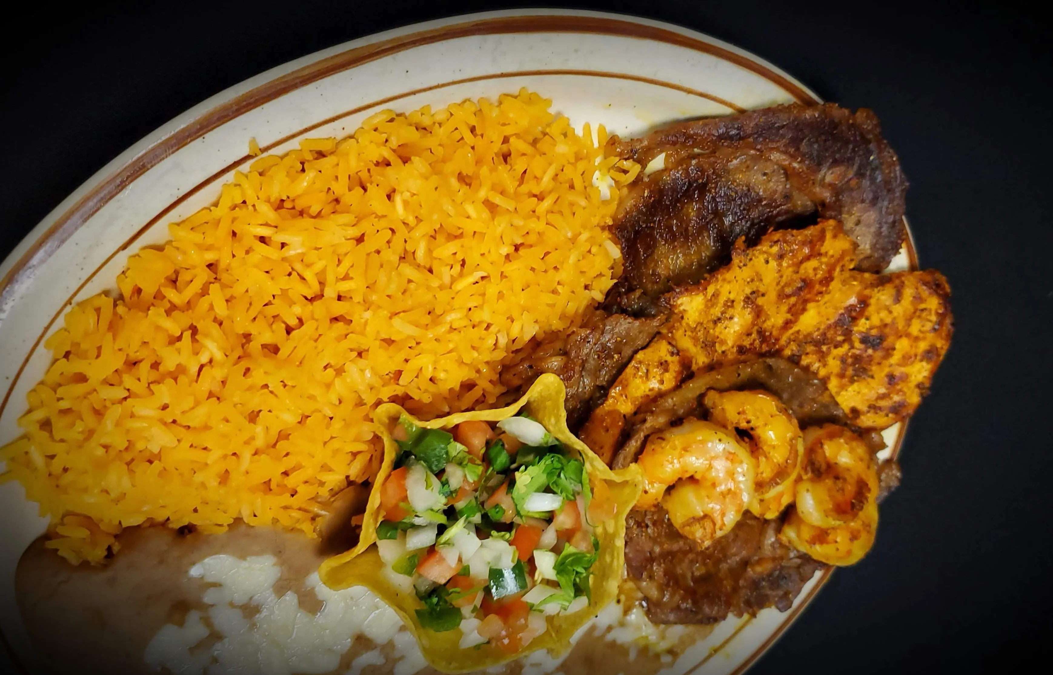 A plate of Tierra Mar y Cielo featuring grilled steak, seasoned grilled chicken, and shrimp, served with Mexican rice, refried beans, and pico de gallo in a crispy tortilla shell.