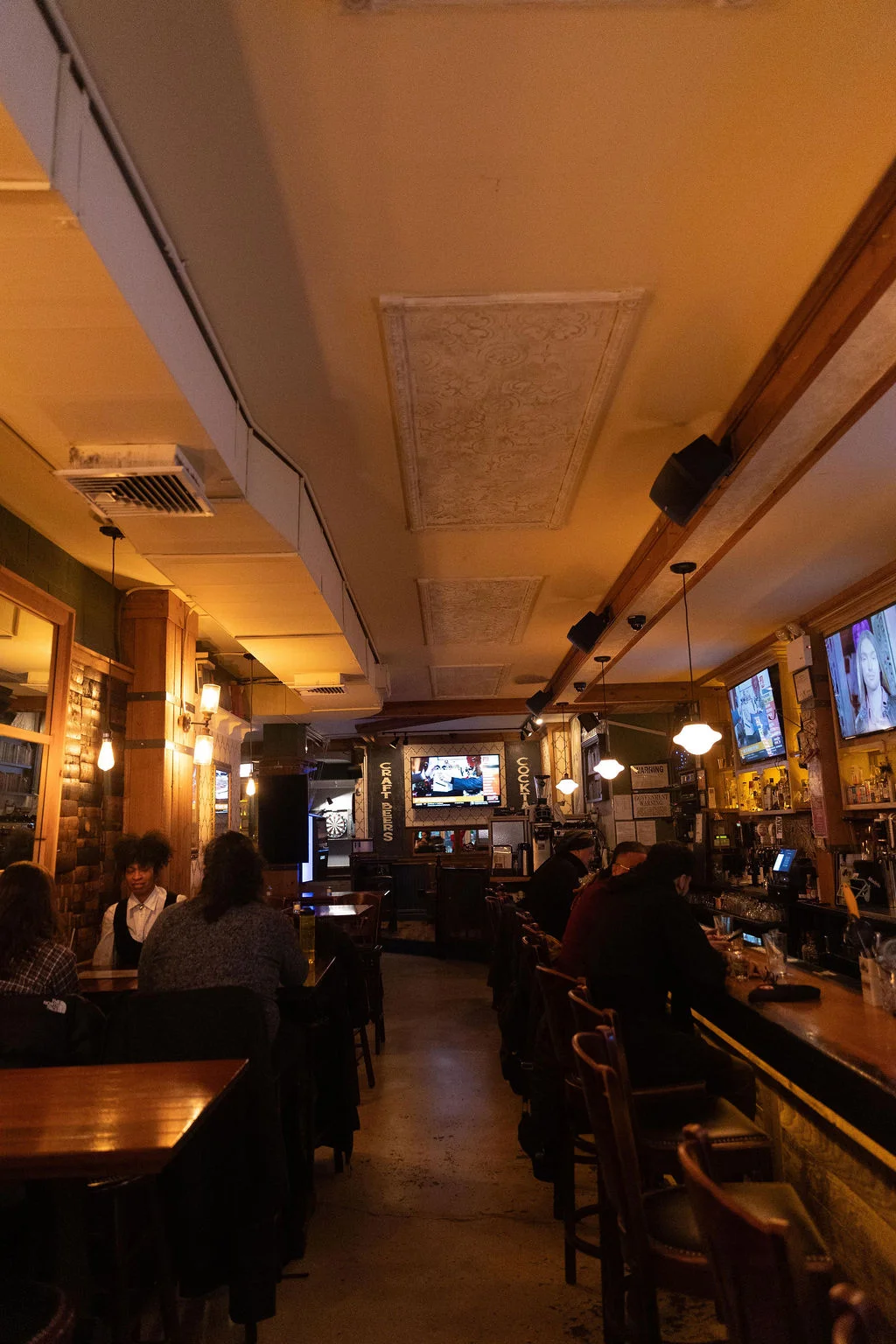 An interior view of a bar, showcasing a long counter, tables, and screens displaying sports. Patrons can be seen seated at tables and the bar.
